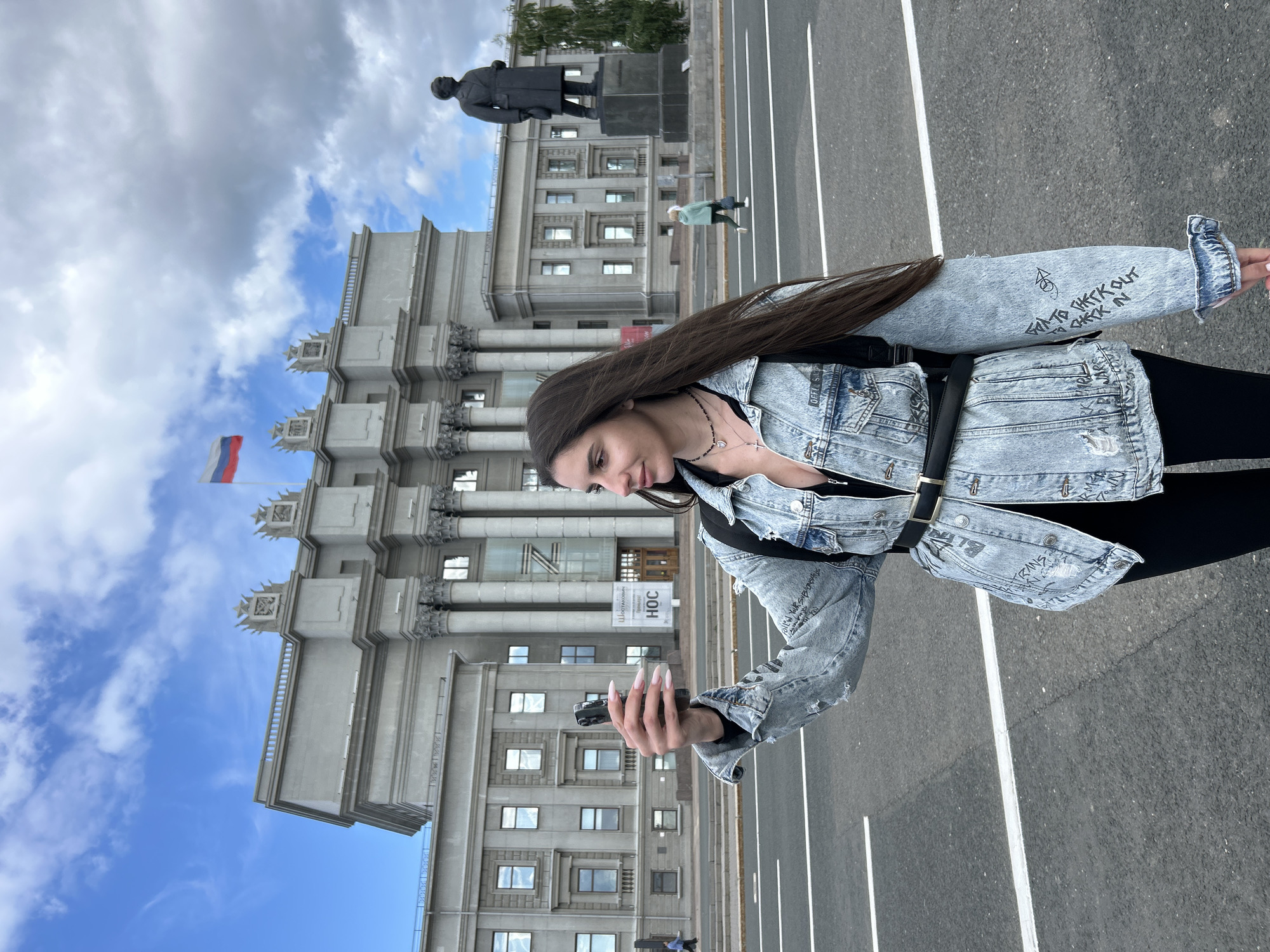 In Novokuybyshevsk, Russia, a young woman with long, dark hair is taking a selfie with her smartphone. She is wearing a light blue denim jacket with distressed details and black writing on the sleeves, a black top, black pants, and a black belt. A black backpack is visible on her shoulders. The background features a large, imposing grey building with columns and a Russian flag flying above it. A statue of a man in a long coat stands to the right of the building. The scene takes place on a street with distinct white lane markings, and in the distance, two pedestrians can be seen. The sky is partly cloudy.