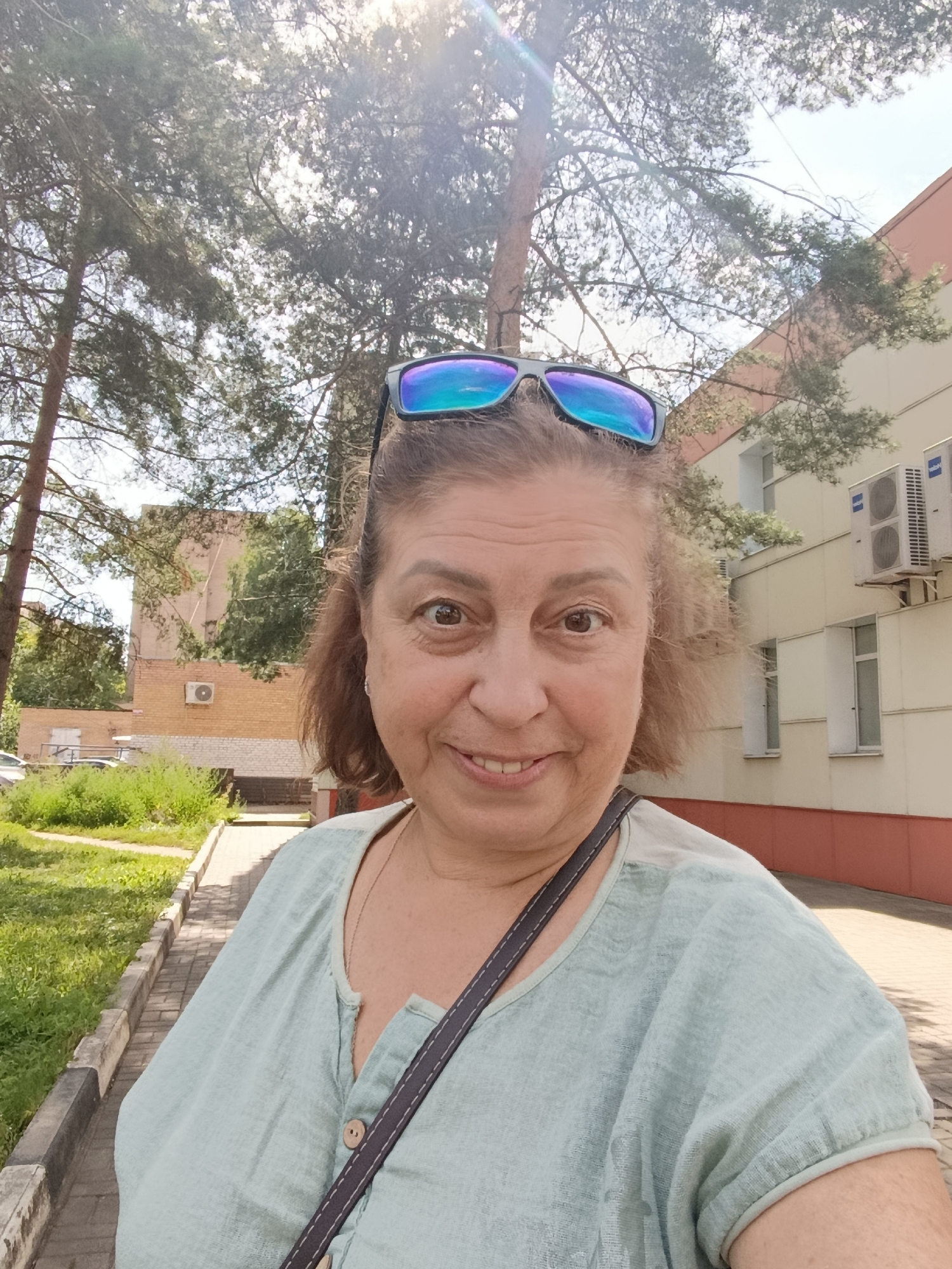 An outdoor, eye-level, medium shot shows a smiling, middle-aged Caucasian woman with brown hair and sunglasses perched on her head, taking a selfie on a sunny day. She is wearing a light blue, short-sleeved blouse with a V-neck and a single button closure. A dark strap of a bag is visible across her chest.
The background features a paved walkway on the right, bordered by a strip of grass and then more grass and bushes on the left. Beyond the walkway, there is a light-colored building with red trim and several air conditioning units mounted on its exterior. A large tree with sparse green foliage is visible to the left of the woman, and another tree is directly behind her head, with its branches and needles filling the upper portion of the frame, obscuring parts of the sky. The overall environment suggests a residential or institutional area within the city of Orekhovo-Zuyevo, Russia, possibly near a park or public space.