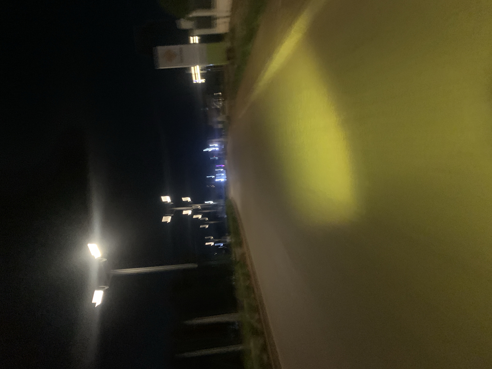 The image depicts a nighttime view of a street in Bukuru, Nigeria. A street light with two lamps illuminates a portion of the asphalt road with a yellow glow. The road is bordered by a sidewalk and vegetation. In the background, several other streetlights are visible, along with a building that has a sign with a yellow emblem. The sky is dark, indicating it is nighttime. The overall scene is blurry, suggesting motion or a low-resolution image.