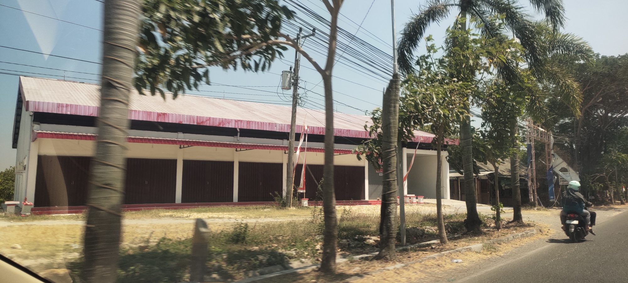 The image shows a commercial building with a red and white striped awning and brown roll-up doors, located in Mranak, Indonesia. A person is riding a motorcycle on the right side of the road, wearing a light blue helmet. The building appears to be a series of storefronts or workshops. Palm trees and other vegetation are visible along the roadside. Electrical wires are strung overhead. The sky is clear and the sun is shining.