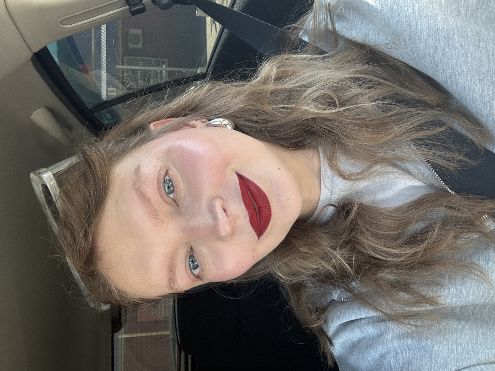 A woman with blonde hair and blue eyes is taking a selfie in a car. She is wearing a grey hoodie and a white t-shirt, and she has on red lipstick and large silver hoop earrings. The background shows the interior of the car and a blurred view of buildings and trees, suggesting the car is on a street in Aksay, Kazakhstan.
