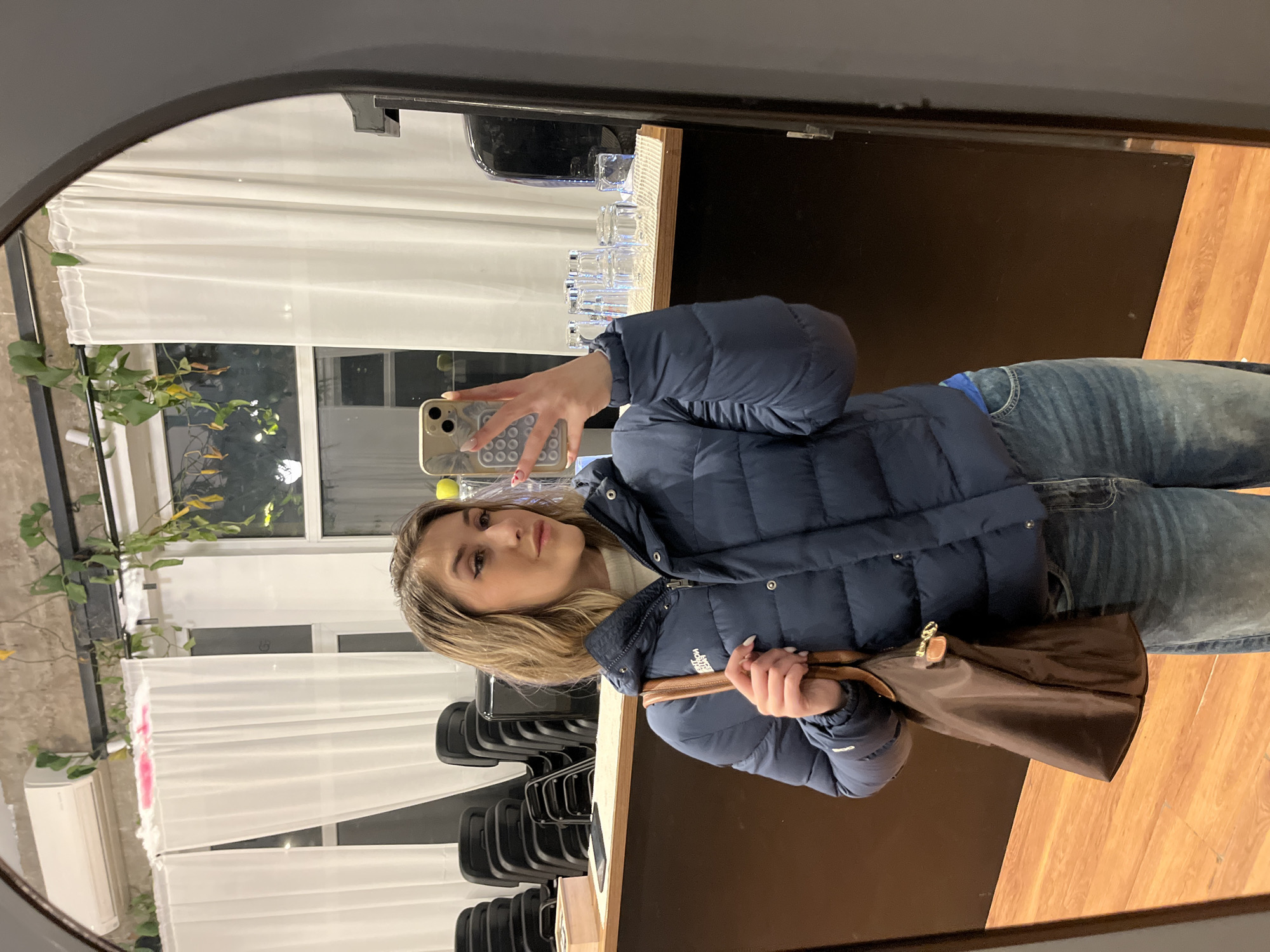 A young woman is taking a selfie in a mirror. She is wearing a blue puffer jacket and jeans, and she is holding a brown bag. The background includes a window with white curtains and plants, a black counter with glasses and stacks of black chairs, and a wood floor. The mirror has a gray frame. The location is not evident in the image.