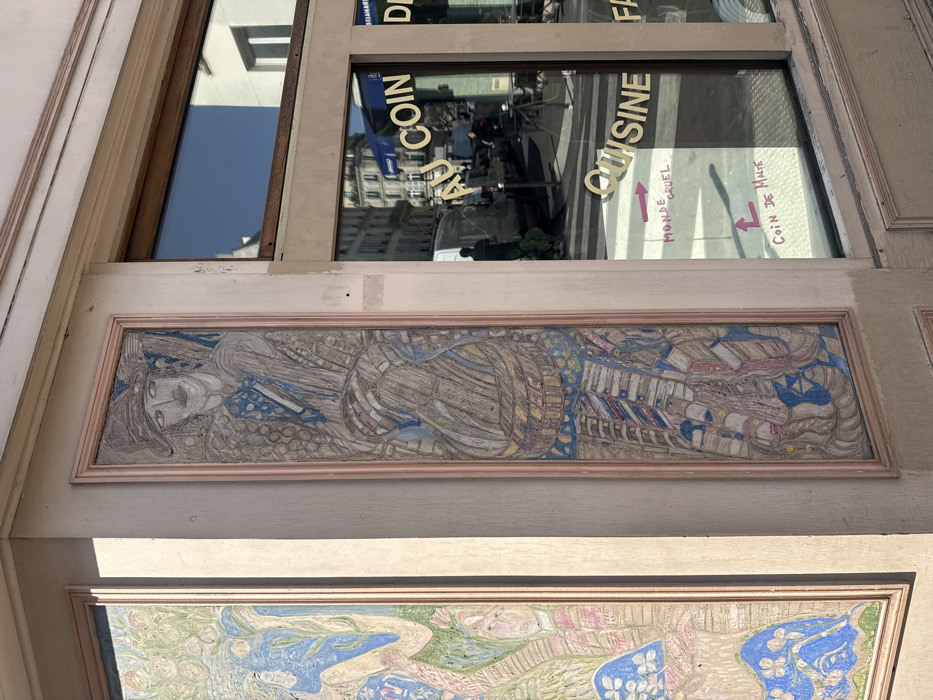 The image displays the exterior corner of a building, identified by signage as "Coin de Malte" (Corner of Malta), indicating a location on Rue de Malte in Paris, France. The scene is illuminated by direct sunlight, casting shadows.
The building facade is light beige, featuring two framed, vertical artworks in a stylized, colorful aesthetic, possibly reliefs or murals. The prominent corner artwork depicts an abstract figure with a distinct face, mustache, and a hat, rendered with blue, grey, and earthy tones, and textured patterns. To its left, a partially visible second artwork presents a figure amidst organic, plant-like forms in blues, greens, and pinks. Both artworks are framed with pale pink trim.
Adjacent to the corner artwork is a large, street-level window with a brown frame. The window glass reflects the urban environment, including typical Parisian Haussmannian architecture, a white delivery van, and a dark-clad individual pedestrian on the street. The upper portion of the window reflects a clear blue sky.
Gold adhesive letters affixed to the window read "AU COIN" and "CUISINE," with additional partial text visible. Below these, handwritten in pink marker directly on the glass, are the phrases "MONDE CRUEL" (Cruel World) with a right-pointing arrow, and "COIN DE MALTE" (Corner of Malta) with an upward-pointing arrow.