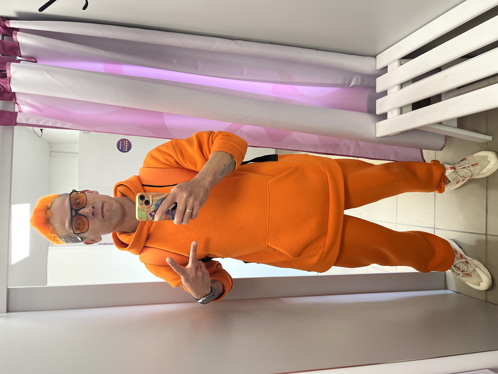A man with orange hair is taking a selfie in what appears to be a dressing room. He is wearing an orange sweatsuit and orange-tinted glasses. He is holding a smartphone with a decorative case. He is also wearing a watch. Behind him is a curtain in shades of white and pink. The dressing room is likely located in Chaikovsky, Russia, although this cannot be confirmed from the image.