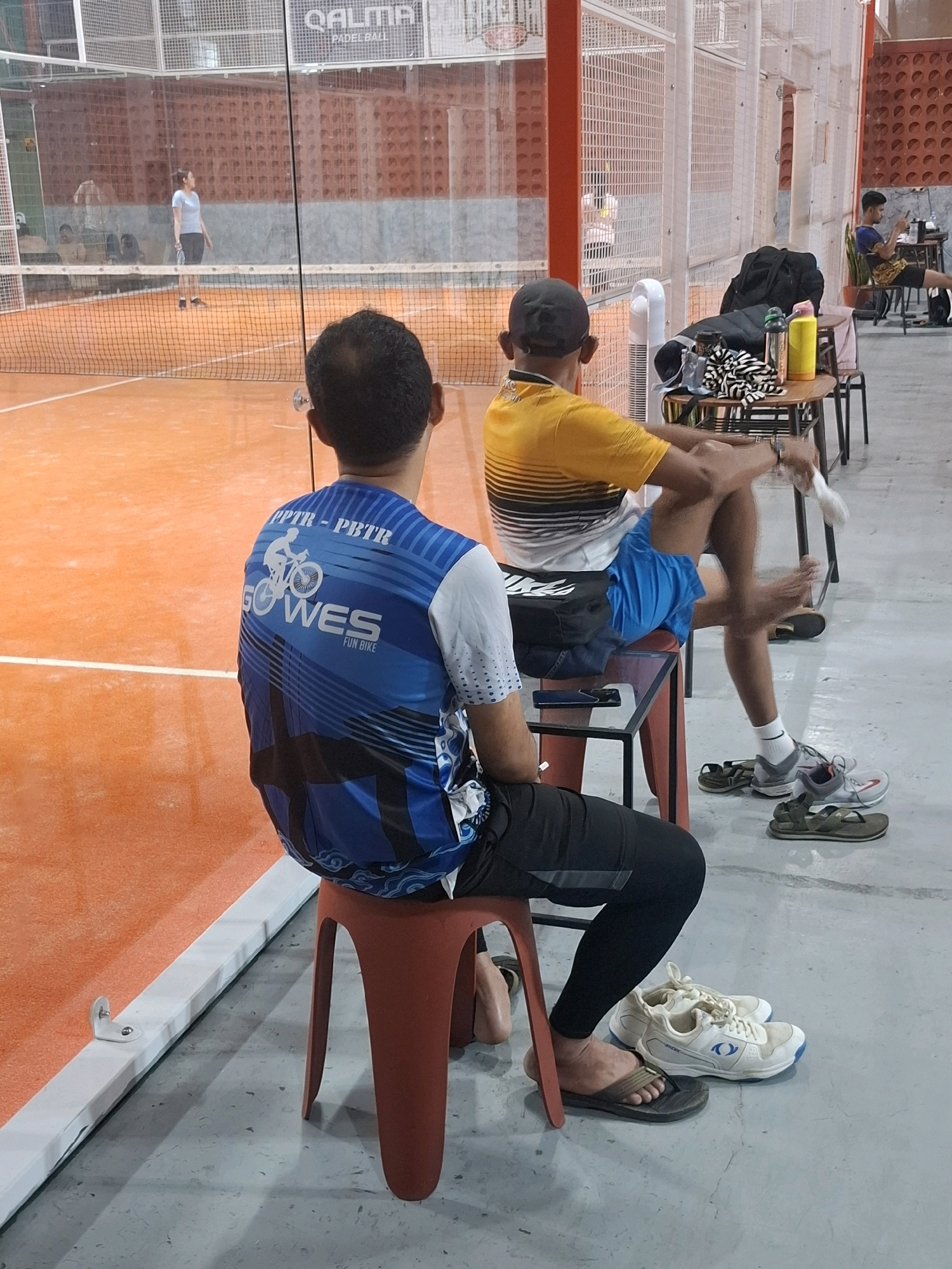 The image depicts an indoor sports facility, likely a padel court, in Cisaat, Indonesia. The court itself has an orange surface and is enclosed by netting and white fencing. In the background, several people are visible. One woman in a light blue top and dark shorts is on the court, holding a racket. Other individuals are seen sitting in the background, some appearing to be spectators or participants resting.
In the foreground, two men are seated on red plastic stools. The man on the left is wearing a blue and white patterned t-shirt with a cycling graphic and black pants. He is facing away from the camera. The man on the right is wearing a yellow and black striped t-shirt and blue shorts. He is sitting with his legs crossed and is holding a white object, possibly a towel or a small cleaning tool, in his right hand. Several pairs of shoes and sandals are scattered on the grey concrete floor around the seated individuals. A small table is placed between them, with various items on it, including bags, water bottles, and what appears to be striped fabric. The overall lighting suggests an indoor environment, possibly with artificial lights supplementing natural light. The setting appears to be casual and recreational.