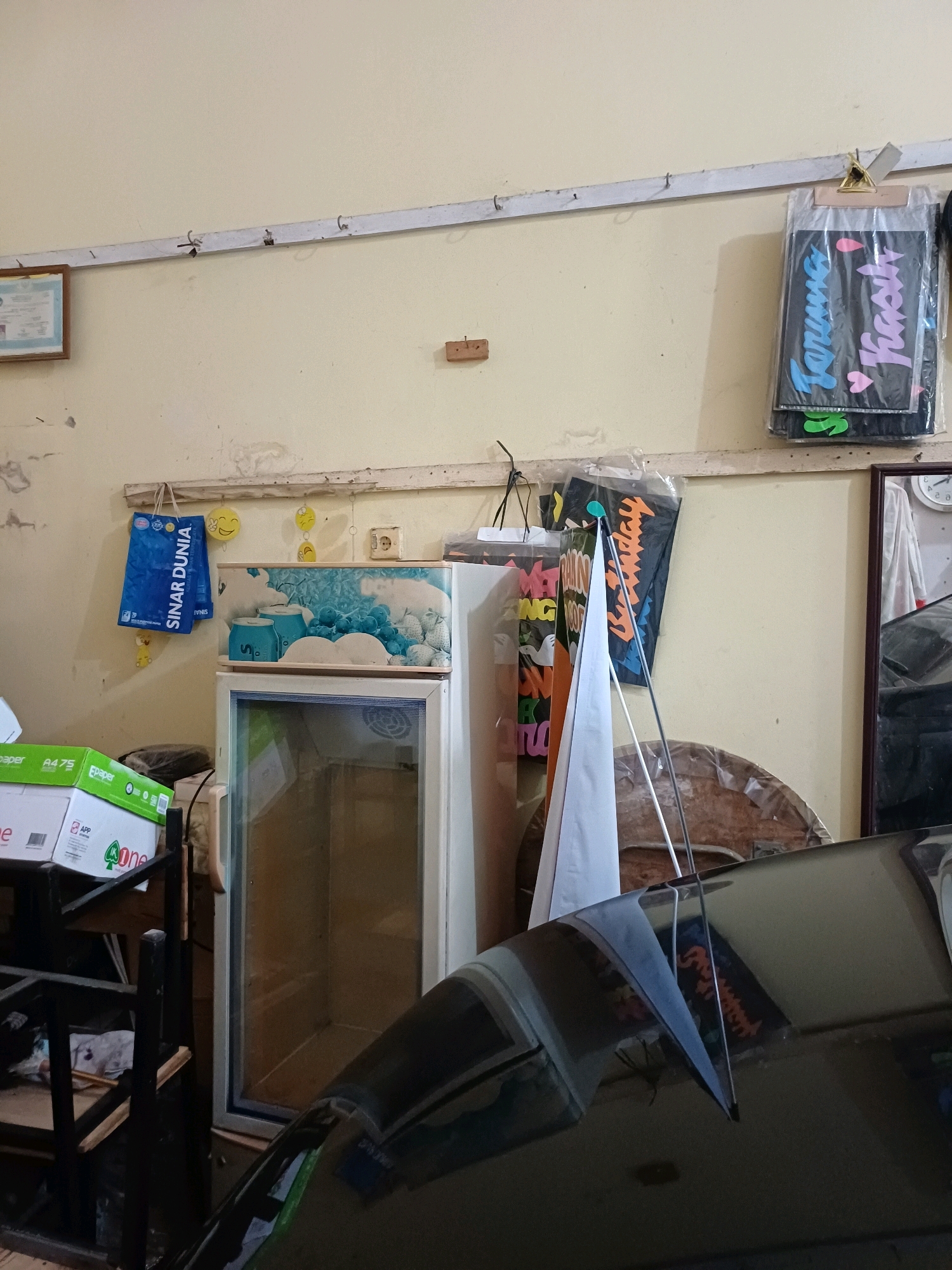 The image depicts a cluttered interior space, likely a shop or storage area, in Dumai City, Indonesia. A light-colored wall forms the backdrop. On the wall, a horizontal metal bar runs across, from which various items are hung.
To the left, a framed certificate or document is affixed to the wall. Below this, a blue shopping bag with the "SINAR DUNIA" logo hangs. Yellow smiley face decorations are also present on the wall.
In the center of the image, a white and blue cooler or refrigerator stands. Above it, various items are displayed on the metal bar, including what appears to be a dark bag with colorful text, possibly signage or decorative elements. One such item has text that appears to read "Terima Kasih" (Thank You in Indonesian). A white flag or banner is also partially visible.
To the right, a framed mirror shows a reflection of a clock on the wall, with Roman numerals. A dark, glossy surface, likely the hood of a vehicle, occupies the lower right portion of the image, reflecting the interior.
In the foreground, to the left, stacks of white boxes with green and blue labeling, possibly paper or office supplies, are visible on a dark metal shelving unit. The overall impression is one of an active but somewhat disorganized commercial or workshop environment.