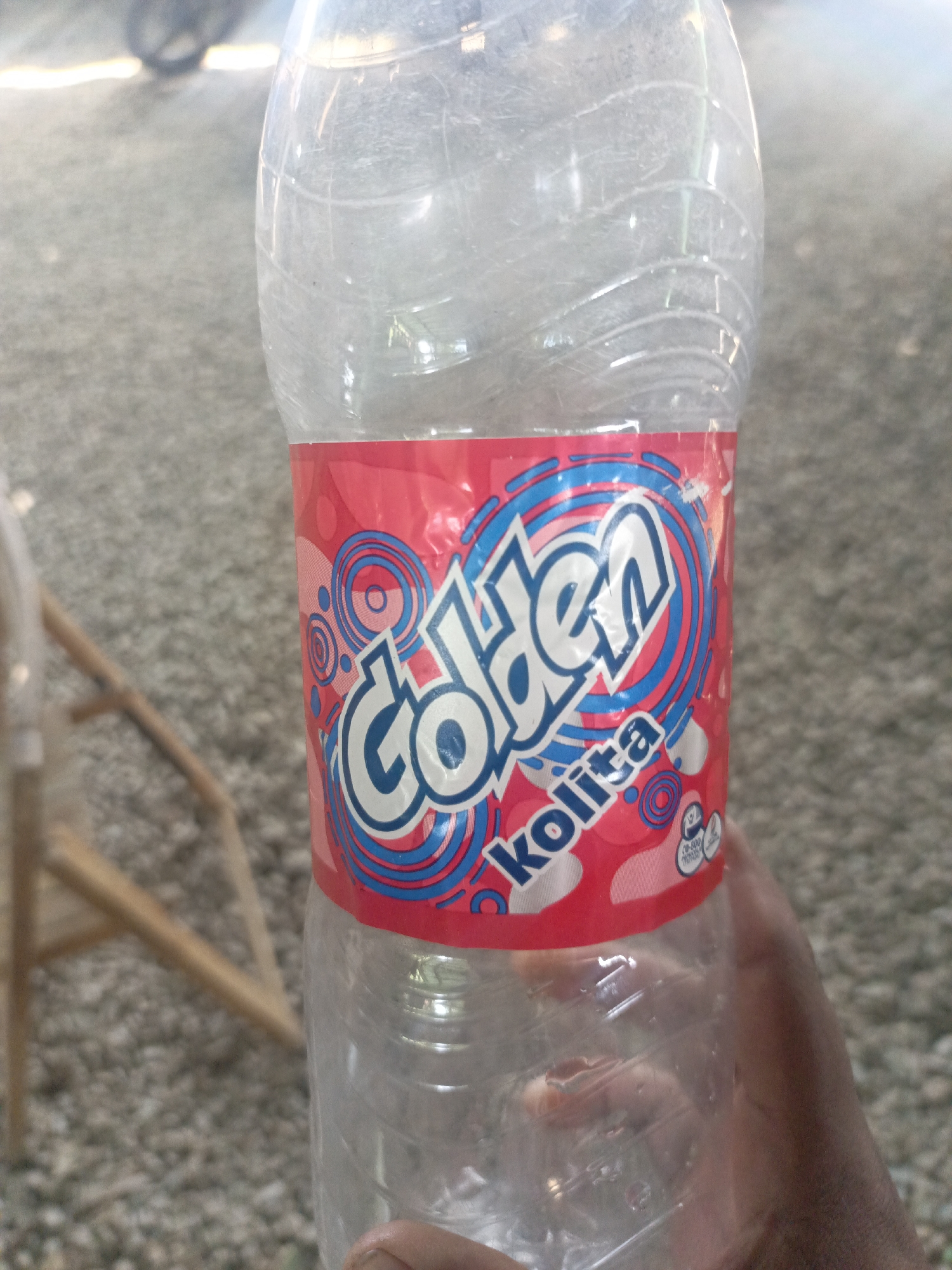 A partially empty, transparent plastic bottle, labeled "Golden Kolita", is held by a person's hand. The background is a gravel-covered area, and the leg of a wooden chair is visible in the lower left. The setting appears to be outdoors. The location is identified as Juan Griego, Venezuela.