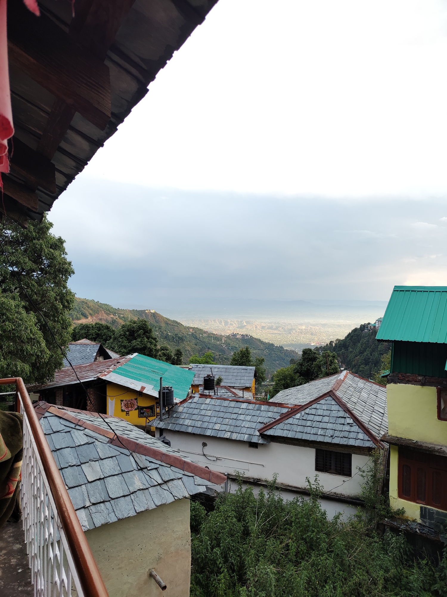 The image presents a view from an elevated position, possibly a balcony, in what appears to be a mountainous region in Haini, India. The foreground is dominated by the roofs of closely packed buildings. The buildings have a mix of materials, including slate shingles on the roofs and a variety of exterior colors such as yellow, white, and green. A white balcony railing is visible. In the background, a city is discernible in the valley, partially obscured by a layer of mist or haze. The horizon is overcast with a grey sky. Green trees and other vegetation are scattered throughout the scene. The lighting suggests it is either early morning or late afternoon. The overall impression is of a densely populated hillside community situated near a larger city in a valley.