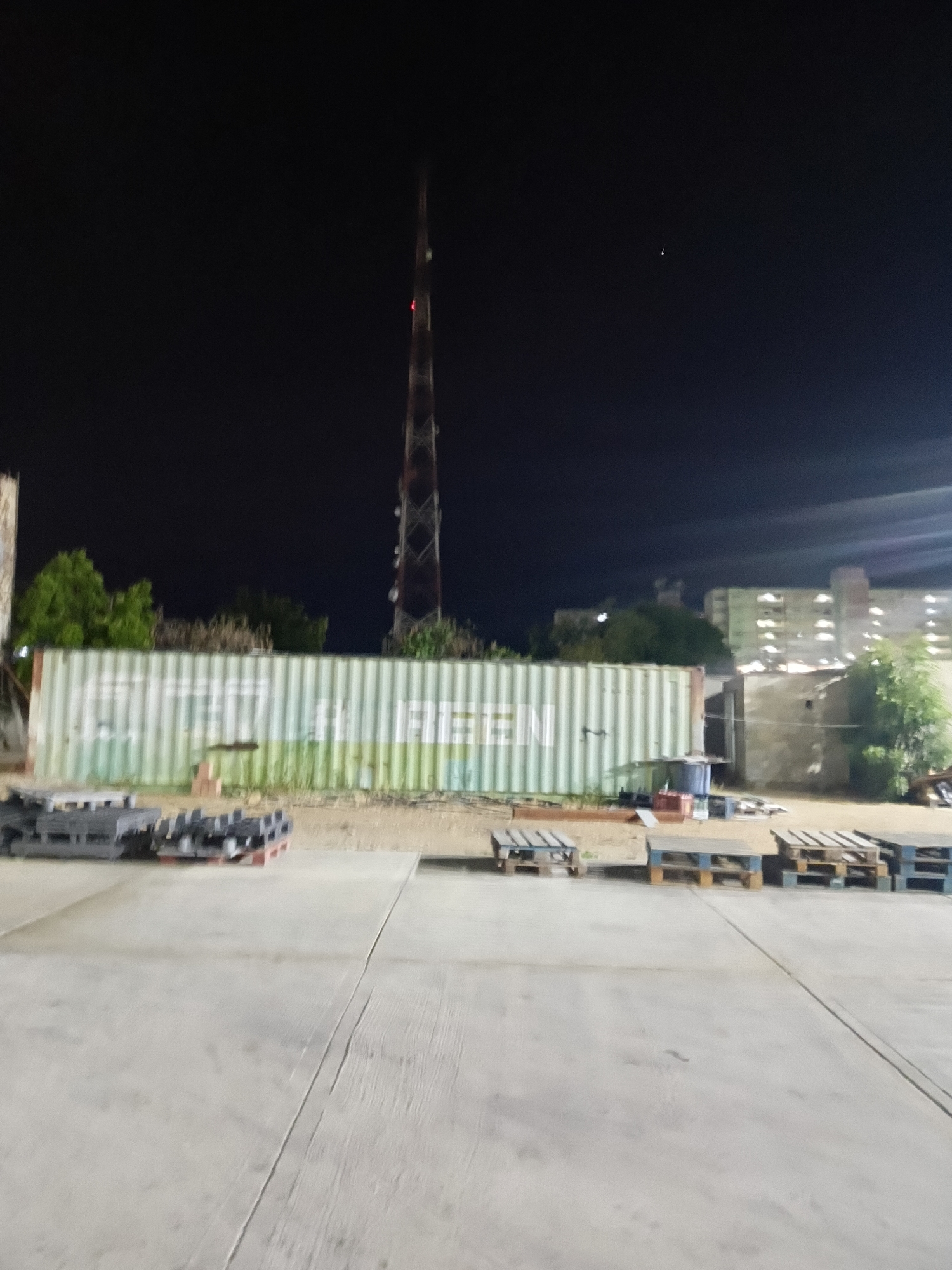 The image, taken at night, shows an open area in Juan Griego, Venezuela. A long, light green shipping container with the word "GREEN" written on it sits in the middle ground. To the container's left are several stacks of dark grey rectangular objects, possibly bricks. Dispersed in front of the container are numerous wooden pallets.
Behind the container, trees and a multi-story building with visible rows of windows can be seen. Further back is a tall, thin, red and grey communications tower extending upwards into the night sky. A small, indistinct point of light, possibly a star or a distant light source, is visible in the dark upper portion of the sky. The foreground is a light grey, paved surface that appears to be concrete or asphalt. There are no people visible in the frame.