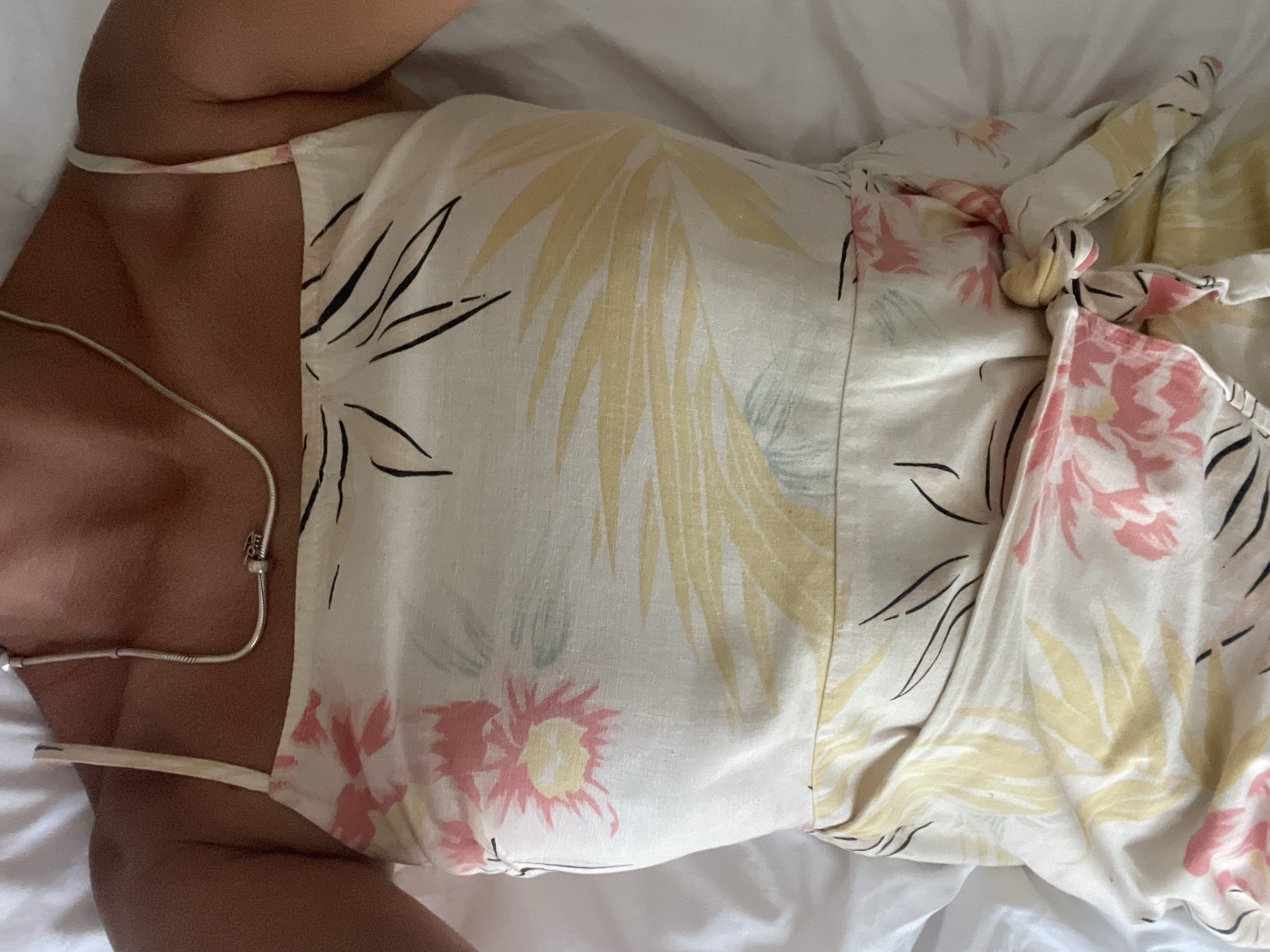 A person is lying on a white sheet, wearing a light-colored dress with a floral pattern, likely indoors. The location is unknown, but the prompt suggests it might be Kuta Selatan, Indonesia. The person is wearing a thin silver necklace. The dress features yellow and pink floral patterns with black leaf outlines. The dress is tied on the side.