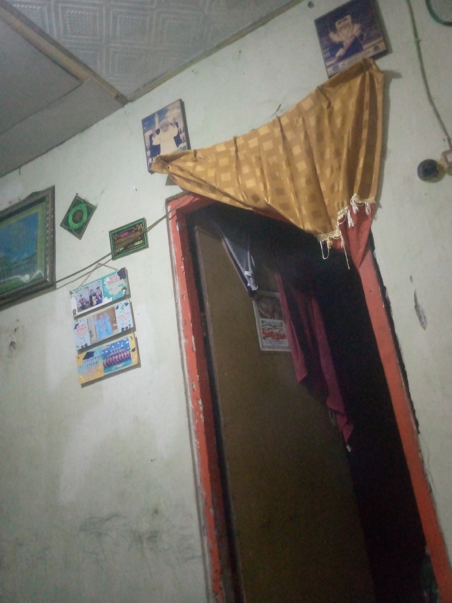 The image depicts an interior scene with a pale, textured ceiling and off-white walls. On the wall, several framed items are visible. In the upper left, a framed picture with a blue and white image, possibly a landscape or building, is hung. Below this is a small, square green frame with an intricate design. To the right of the green frame, a rectangular green frame with Arabic script is mounted. To the right of that, a small framed picture of a person in traditional attire is present. Hanging below the green frames is a vertical display of three rectangular items, each containing images and text, likely photographs or certificates. These are suspended by strings. A doorway is visible on the right side of the image, partially covered by a yellow and gold patterned curtain with decorative fringe. The doorway's frame is painted a reddish-orange color. The environment appears to be a domestic setting.
