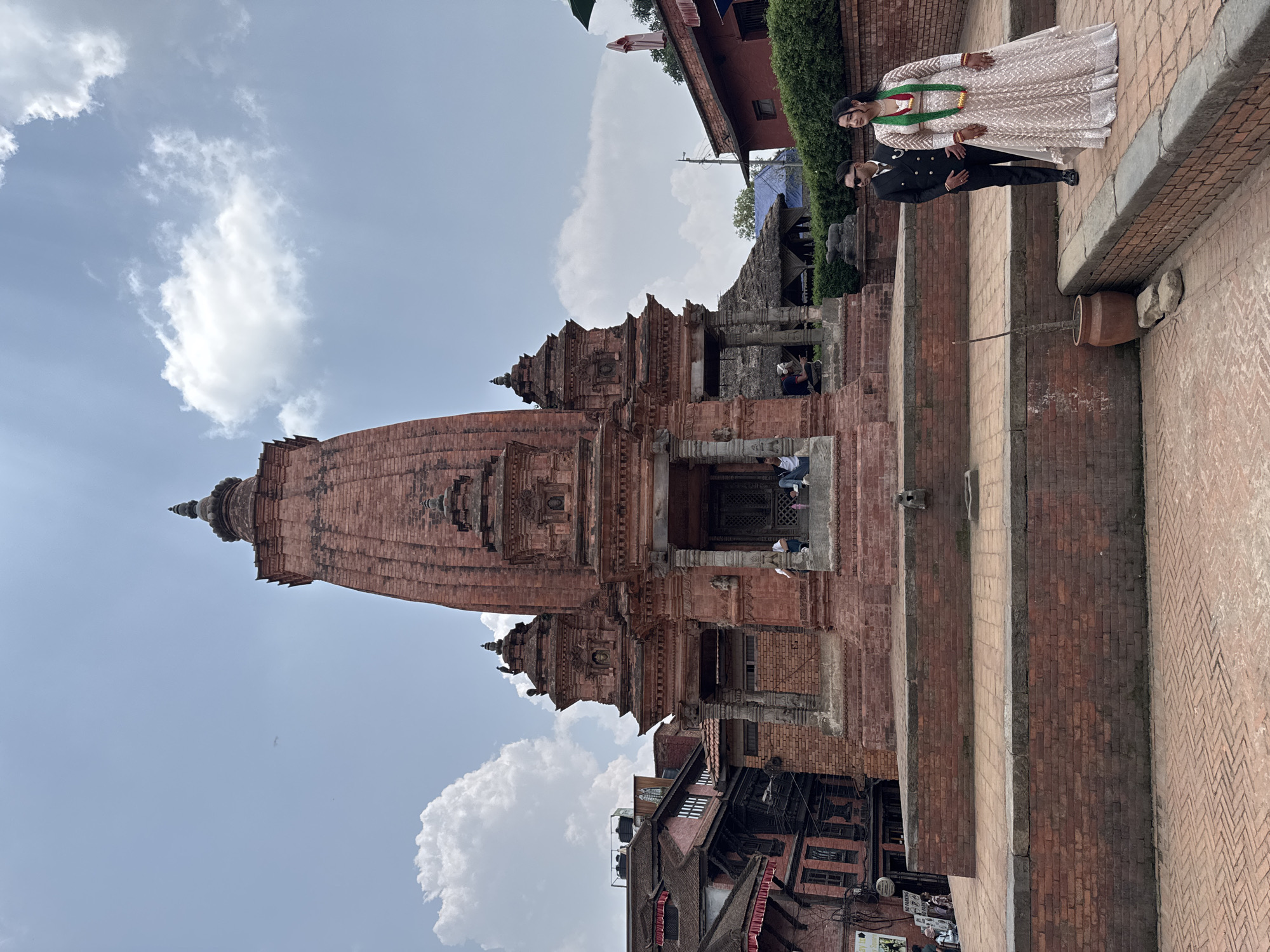 A photograph shows a man and woman standing on the steps of a brick building, with a large brick temple structure in the background and a cloudy sky above, possibly in Bhaktapur, Nepal. The woman is wearing a light-colored, ornate dress with a green garland, while the man is in a black suit and sunglasses. The temple has multiple levels and detailed carvings, and is framed by clouds in the sky. Older buildings are visible at the base of the temple, showing signs of aging with exposed wiring. The steps in the foreground are made of the same reddish-brown brick as the temple.