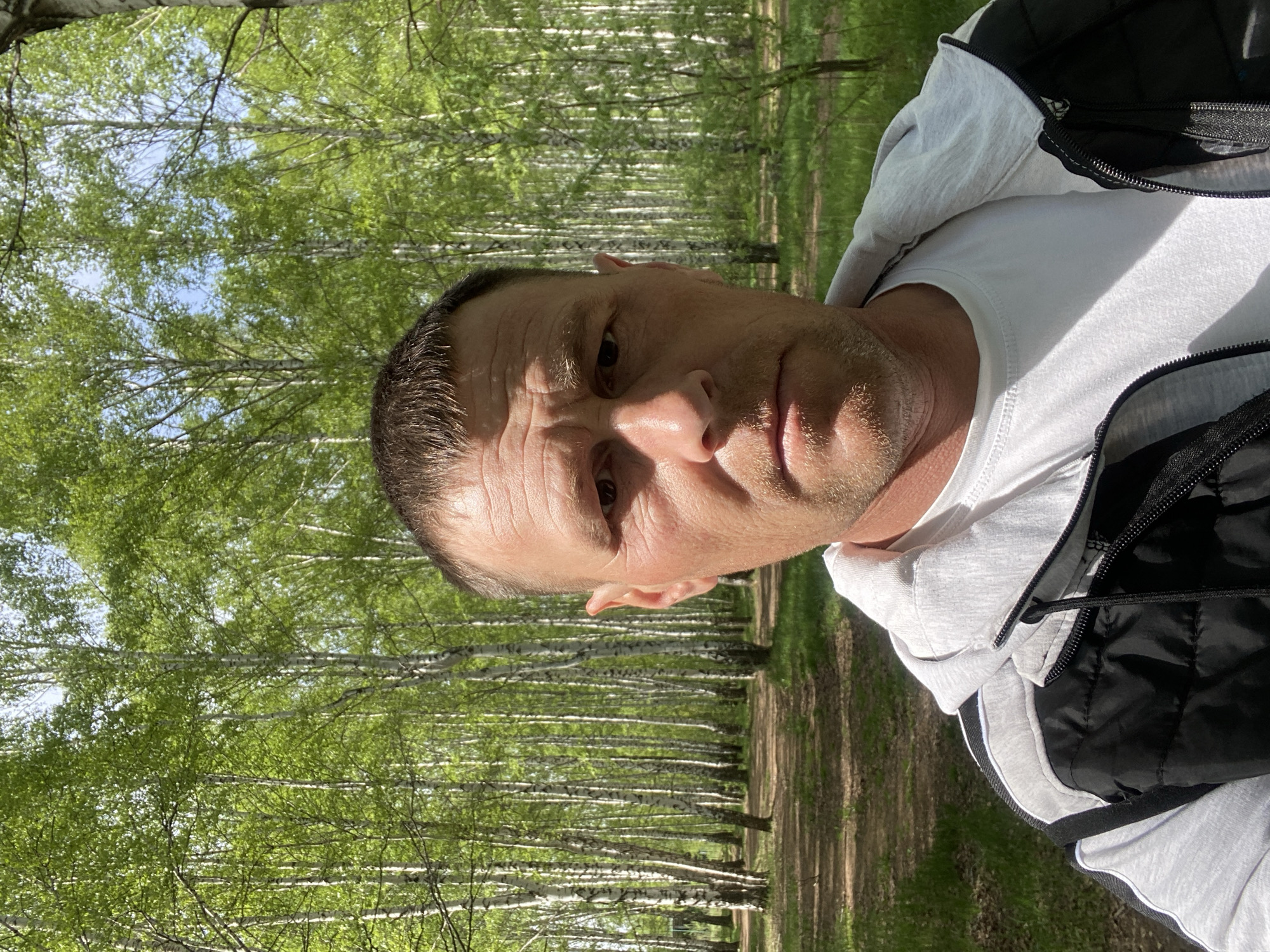 The image shows a man in a white T-shirt, light grey hoodie and black puffer vest, possibly taking a selfie. He is outdoors in what appears to be a birch forest. The background consists of rows of birch trees with green foliage and sunlight filtering through the leaves. The ground is covered in grass and dirt. The location is consistent with a wooded area within the city of Chaikovsky, Russia.