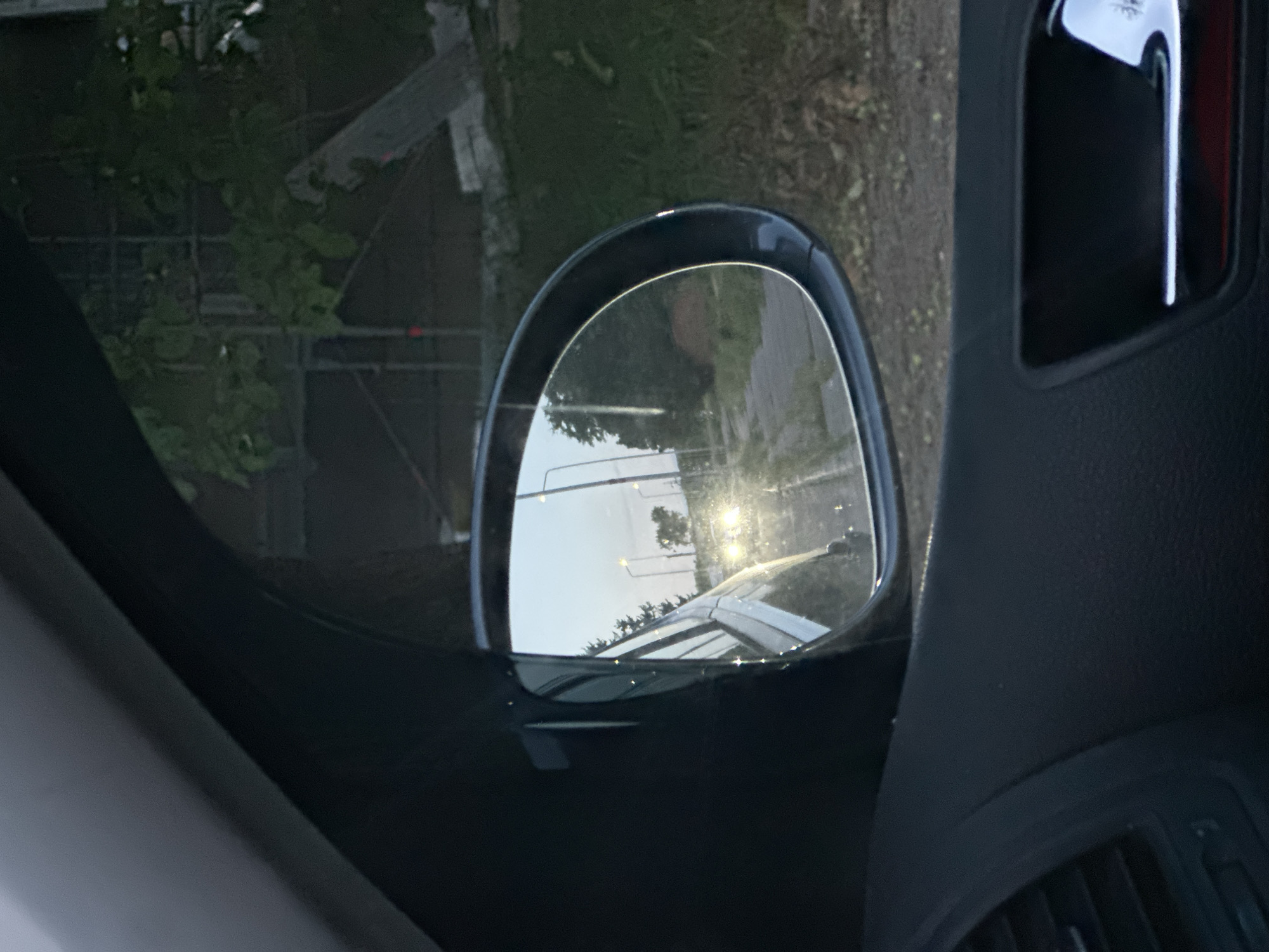 A car's side mirror is the primary subject, reflecting a scene from Lovech, Bulgaria, judging by the architecture, road layout, and street lighting visible in the mirror's reflection. The mirror's reflection shows a road with streetlights, with a car behind the vehicle from which the image is taken. The car's panel, door, and vent can be seen surrounding the mirror.