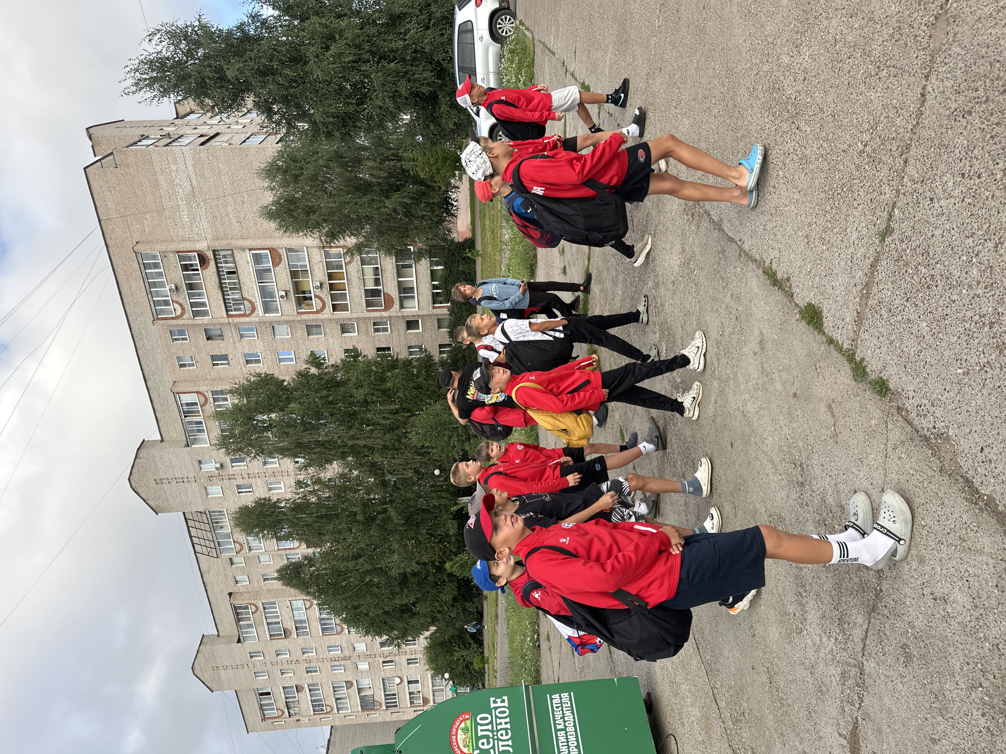 In the city of Chaikovsky, Russia, a group of young boys, likely athletes, are walking outdoors on a paved surface. They are dressed in athletic attire, with many wearing red jackets and shorts. Most of the boys are carrying backpacks. They are walking in front of a multi-story brick apartment building, with trees lining the sidewalk. A large green advertisement sign is visible on the left side of the image. The sky is overcast, indicating a cloudy day. The overall scene depicts a group of boys on the move in a residential urban environment.
