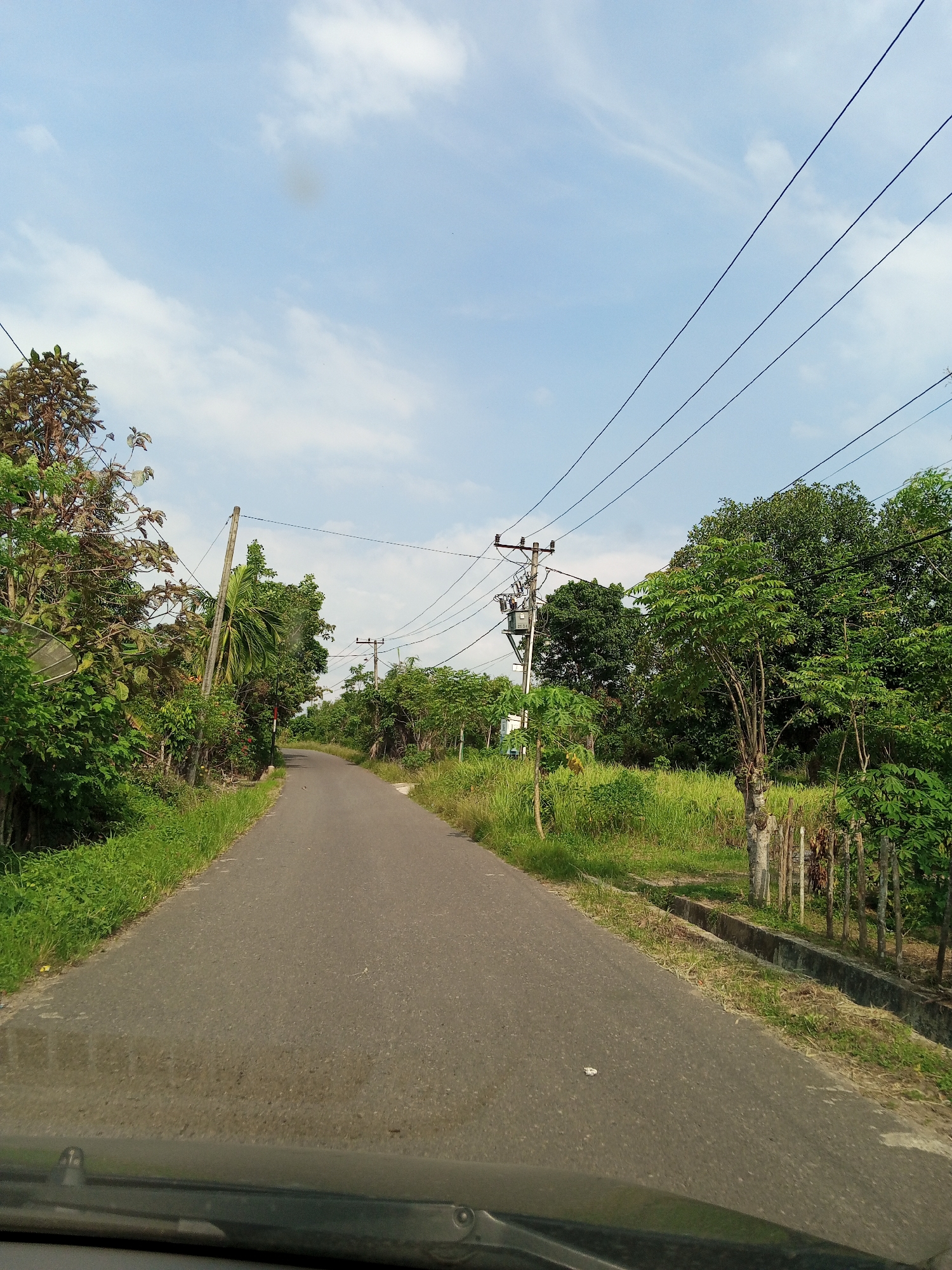 The image depicts a narrow asphalt road in Keudesimpangempat, Indonesia. The road curves slightly ahead and is flanked by dense vegetation on both sides. Utility poles with power lines run along the roadside. The sky is partially cloudy. The view is from inside a vehicle, as evidenced by the dashboard visible at the bottom of the frame. There are no people visible. The environment suggests a rural or semi-rural location.