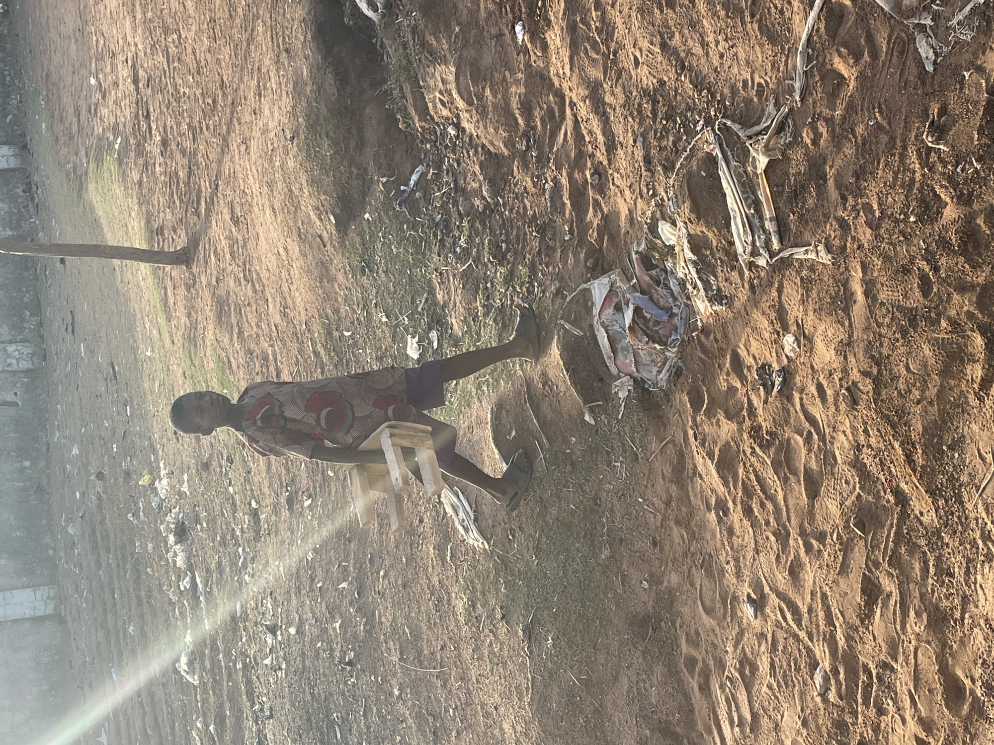 The image depicts a young person carrying a small, wooden chair in Akwanga, Nigeria. The person is walking across a dirt surface littered with trash and debris. The environment appears arid and degraded.