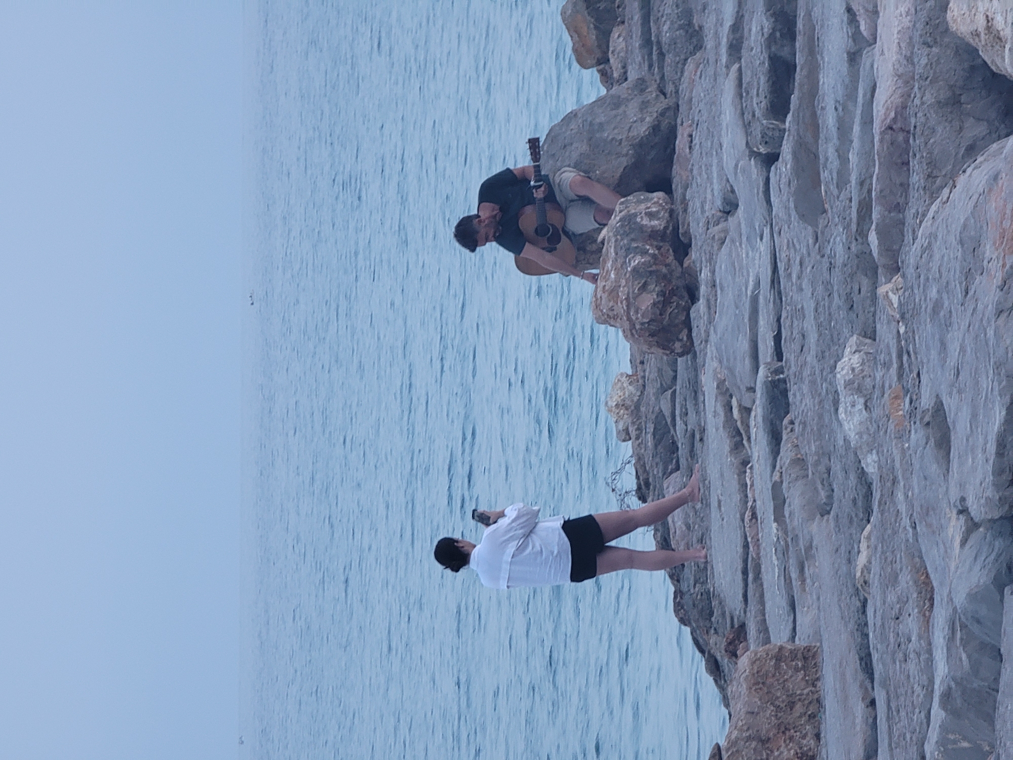In Quarteira, Portugal, a man sits on a large rock on a stone jetty, playing an acoustic guitar. A woman stands on the jetty a short distance away, facing the man and holding a smartphone, as if taking a picture or video. The sea is calm and blue in the background, with a hazy sky. The jetty is made of irregular grey stones.