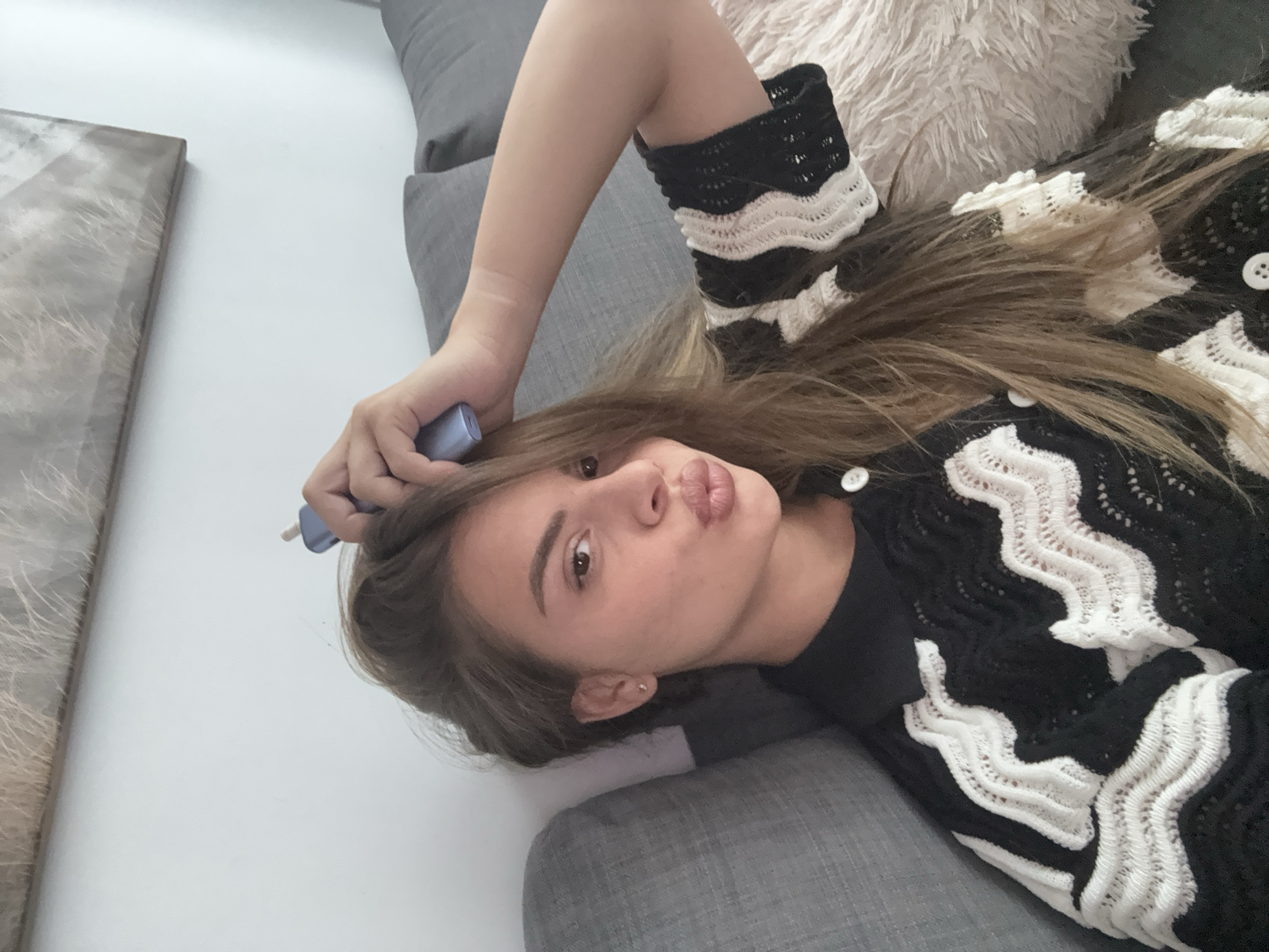 A young woman with long, light brown hair is pictured lying on a grey sofa, looking directly at the camera with a playful pout. Her head is positioned towards the upper right of the frame, and she is wearing a distinctive black and white knitted cardigan or sweater. The cardigan features wavy white patterns against a black background, with visible white buttons. Underneath, she appears to be wearing a plain black top.
In her right hand, which is resting near her head, she holds a small, light blue or purple cylindrical object, resembling an electronic vape pen or similar device. Behind her head, a fluffy, light-colored (possibly cream or white) pillow is visible. To the left side of the frame, a rectangular object, likely a framed picture or canvas art with muted brown and grey textures, leans against a plain white or off-white wall. The overall setting appears to be a comfortable indoor domestic space, such as a living room or bedroom. The woman's expression conveys a confident and playful mood. There is no visible text in the image.