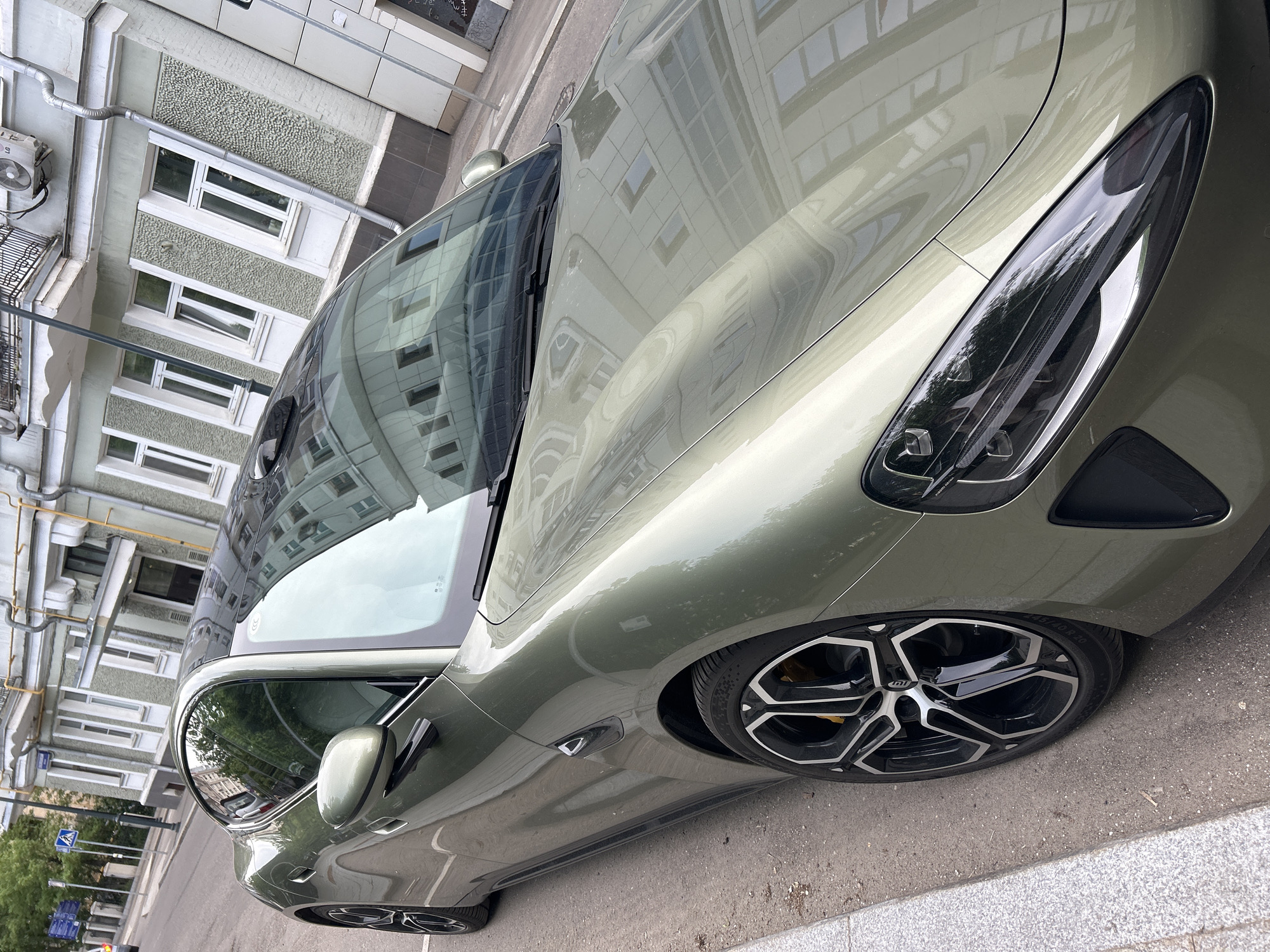 A high-angle view captures a green Kia Stinger parked on a city street, with the rear of the car facing towards the top right of the frame. The vehicle is positioned on a gray asphalt road with a light gray concrete curb visible along the bottom right edge. The wheels are black with silver accents.
Reflections of buildings are visible on the car's surface. Behind the car, there's a multi-story building with white-framed windows and an architectural design of vertical panels with a gray speckled surface. The buildings further in the background are reflecting in the car's paint.
The image suggests an urban setting with the car parked alongside a typical city street. The location is labeled as Unknown, Unknown. There are no people in the frame.