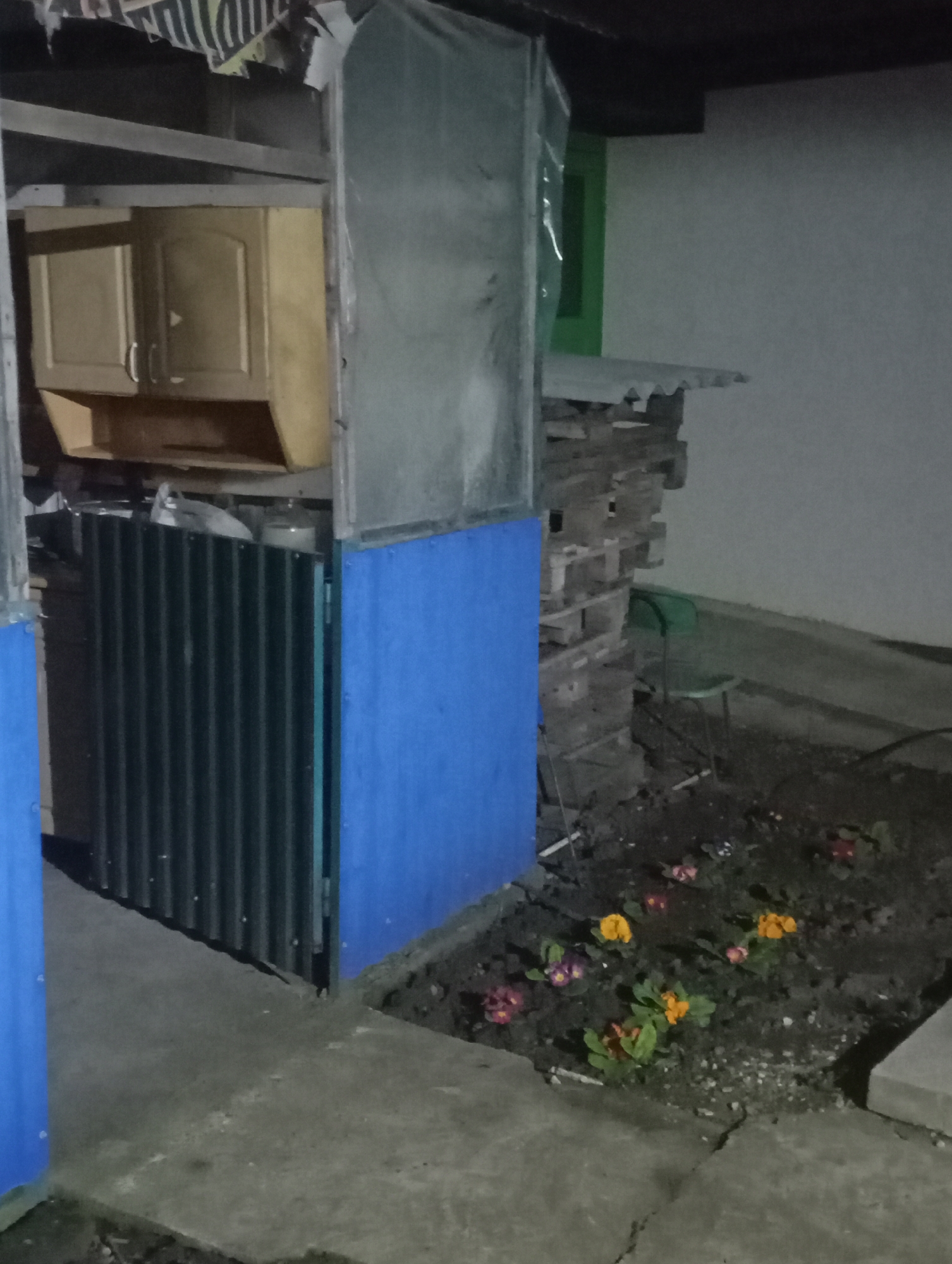 The image depicts an outdoor scene in Kurganinsk, Russia, at what appears to be dusk or night due to the dim lighting. The foreground features a cracked concrete surface on the left leading to a dark soil bed on the right, containing approximately ten small flowering plants with yellow, orange, red, and purple blossoms. A concrete block is visible at the far right edge of the soil bed.
On the left side of the scene, a makeshift structure is present. This structure includes vertical blue panels in the lower left, appearing to be part of an enclosure. Above these, a dark corrugated metal sheet stands vertically. Behind this sheet, a white plastic bag and a light-colored gas cylinder are partially visible. Above this assembly, a light brown wooden wall cabinet with two doors (one slightly ajar) and an open lower shelf is suspended from an unseen support.
Towards the center-right, a tall, vertical silver-grey panel is positioned. To its right, a plain white wall is visible, featuring a distinct green window frame in the upper background. Below the silver-grey panel and against the white wall, a stack of wooden pallets forms a rough wall, topped by a light grey corrugated sheet serving as an overhang. A green plastic chair is situated behind this stack of pallets. The overhead area of the scene shows a dark ceiling or roof structure with torn paper or fabric remnants hanging down. No people or animals are present, and no actions or interactions are depicted.