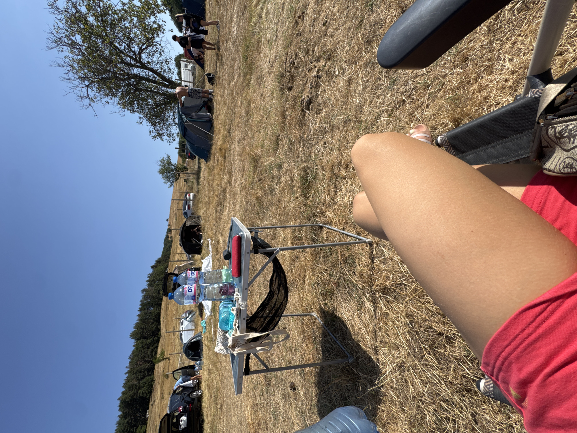 The image depicts a campsite on a dry, grassy field, likely in or near Zheravna, Bulgaria. Several tents are pitched, and vehicles are parked in the background, suggesting a festival or outdoor gathering. A portable table is set up with two bottles of water and a blue plastic bottle. In the foreground, a person is seated in a chair, with their legs visible. The scene is under a clear blue sky, indicating a sunny day. The landscape suggests a rural or hilly area with trees in the distance.