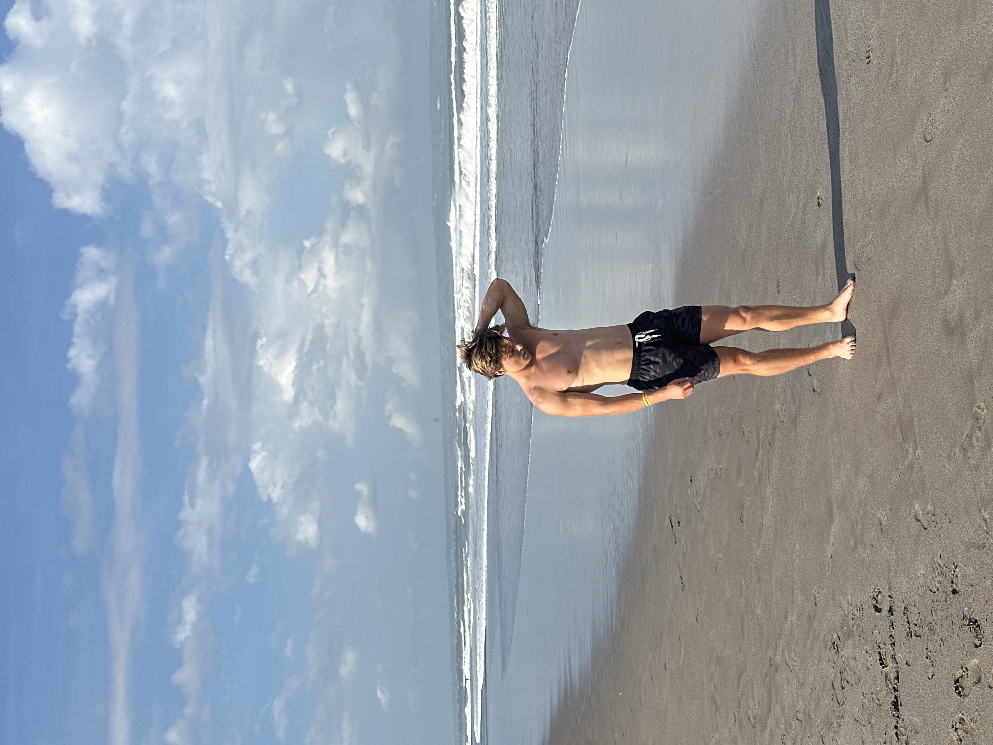 A young Caucasian male with blonde hair stands on a sandy beach in North Kuta, Indonesia. He is wearing black patterned swim trunks with a white drawstring and a yellow bracelet on his left wrist. His left arm is raised, with his hand in his hair. The ocean is to his right, with waves breaking on the shore. The sky is blue with scattered white clouds. The sand is wet near the water, with footprints visible. In the distance, there are hills and possibly some buildings.