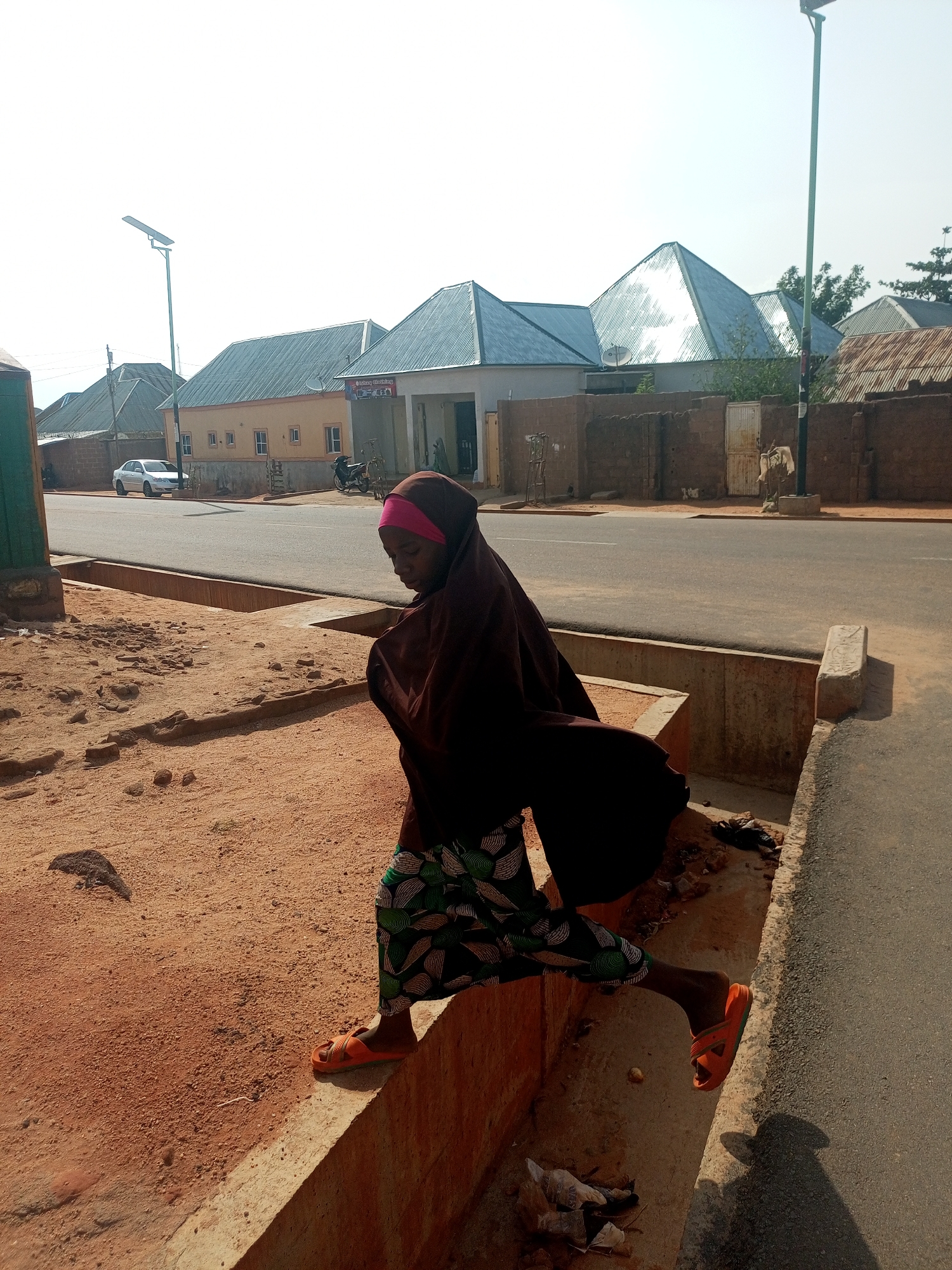 A woman wearing a brown hijab, a pink headscarf, and a green patterned skirt is jumping over a concrete ditch on a street in Tempure, Nigeria. She is wearing orange sandals. The street is paved and bordered by buildings with corrugated metal roofs and brick walls. A white car is parked on the side of the street in the background. A street light is also visible. The ground is reddish-brown dirt. Debris and trash are visible in the ditch.