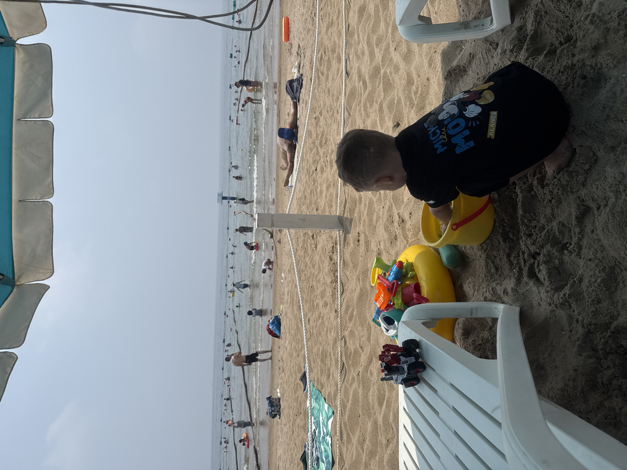 A wide shot shows a sandy beach with people swimming and playing in the ocean under a bright, hazy sky. A blue and white beach umbrella shades the foreground. In the mid-ground, a man lies prone on the sand. In the foreground, a young child in a black t-shirt with a Mickey Mouse design is playing in the sand with a yellow bucket and toy construction vehicles. The beach is located at a resort in Qinhuangdao, China.