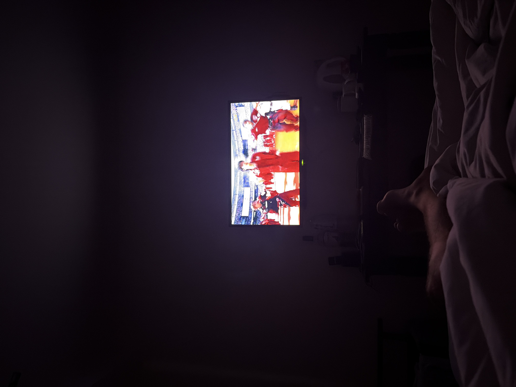 In a dimly lit room, a television screen displays a group of individuals in red attire, seemingly performing on a stage or in a stadium setting. The television is positioned above a dark entertainment unit. To the right of the unit, a white object, possibly a speaker or a fan, is visible. In the foreground, a person's leg, covered in hair, is resting on a white comforter, suggesting they are in bed. The overall ambiance is dark, with the television providing the primary light source. The scene appears to be captured from a low angle, looking upwards towards the television.