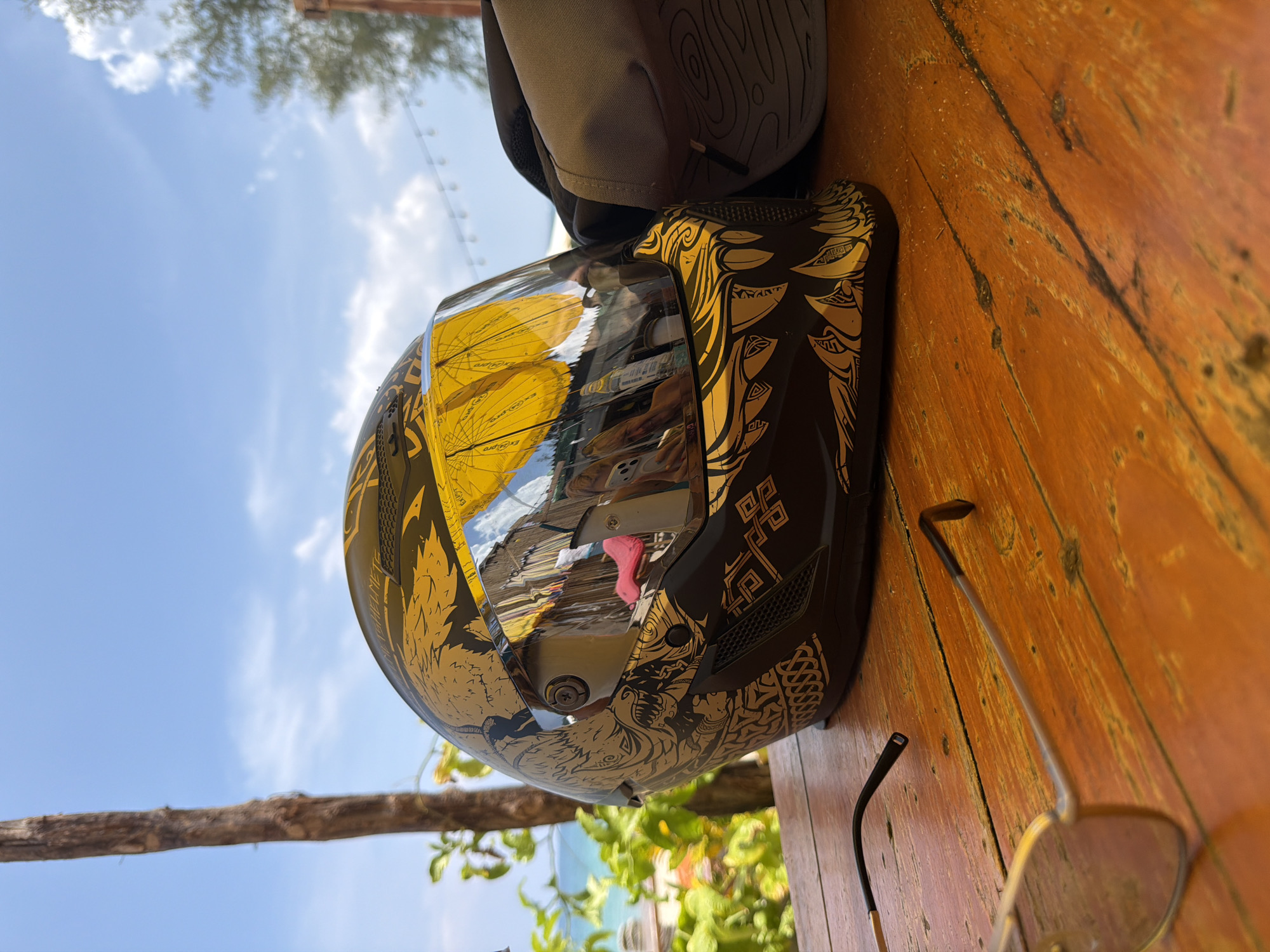 An outdoor, brightly lit scene captures a black and gold motorcycle helmet resting on a rustic wooden table. The helmet, positioned on its side, features intricate golden patterns resembling Celtic knots, mythical beasts, and abstract designs against its dark matte finish. Its clear visor reflects the vibrant surroundings: yellow beach umbrellas, distant buildings, a clear blue sky with scattered white clouds, and hints of green foliage. A prominent reflection on the visor includes partial text that appears to be "EXP_RO".
Next to the helmet, a gray fabric backpack or bag is visible, featuring a subtle, swirled pattern on its top surface. Partially visible sunglasses with metallic frames lie on the wooden table, which has a reddish-brown hue and shows distinct grain and wear.
The background reveals a sunny day with a bright blue sky and sparse white clouds. Green leafy trees are visible, particularly in the lower left, suggesting a lush, possibly tropical, environment. A natural wooden branch extends across the middle left of the frame. The reflections in the helmet visor, particularly the beach umbrellas and coastal elements, strongly suggest a location near a beach or resort. The overall setting implies a pause or break, with personal items placed on a table in an open-air environment, consistent with a location like Choeng Thale, Thailand. The time of day is clearly daytime, given the bright sunlight. No direct human activity is depicted, but the arrangement of objects suggests recent use or anticipation of use.