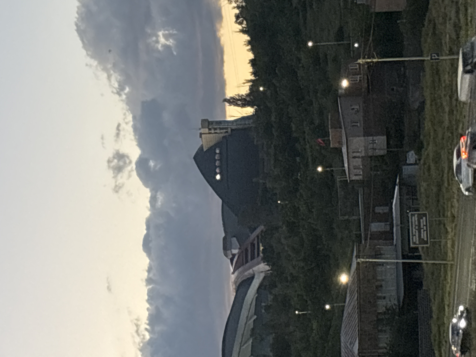 The image captures an evening or dusk scene in Yerevan, Armenia, characterized by a dramatic sky and urban landscape. The sky, positioned on the left when viewed upright, features significant dark, heavy cloud cover. However, a prominent band of golden-orange light stretches across the horizon, indicating either a sunset or sunrise, with the presence of artificial lights suggesting evening.
The central subject is a large, dark, and architecturally distinctive building. Its design is modern, with an angular or curved profile that could be interpreted as ship-like or futuristic. Several circular, illuminated windows or decorative lights are visible along its facade. This structure is set against a backdrop of densely treed hills or mountains.
On the right side of the image, representing ground level, an urban environment is visible. Multiple streetlights are illuminated, casting bright spots of light. Sections of a road or highway can be seen, along which several cars are traveling, their headlights or taillights glowing. Interspersed among the trees and along the roadside are smaller, more conventional buildings. A dark, rectangular sign is mounted on a pole near the road, but any text on it is too indistinct and blurry to be legible.
No people are visible within the frame. The overall atmosphere is calm and transitions from day to night, highlighted by the interplay of the fading natural light and the newly active artificial illumination.