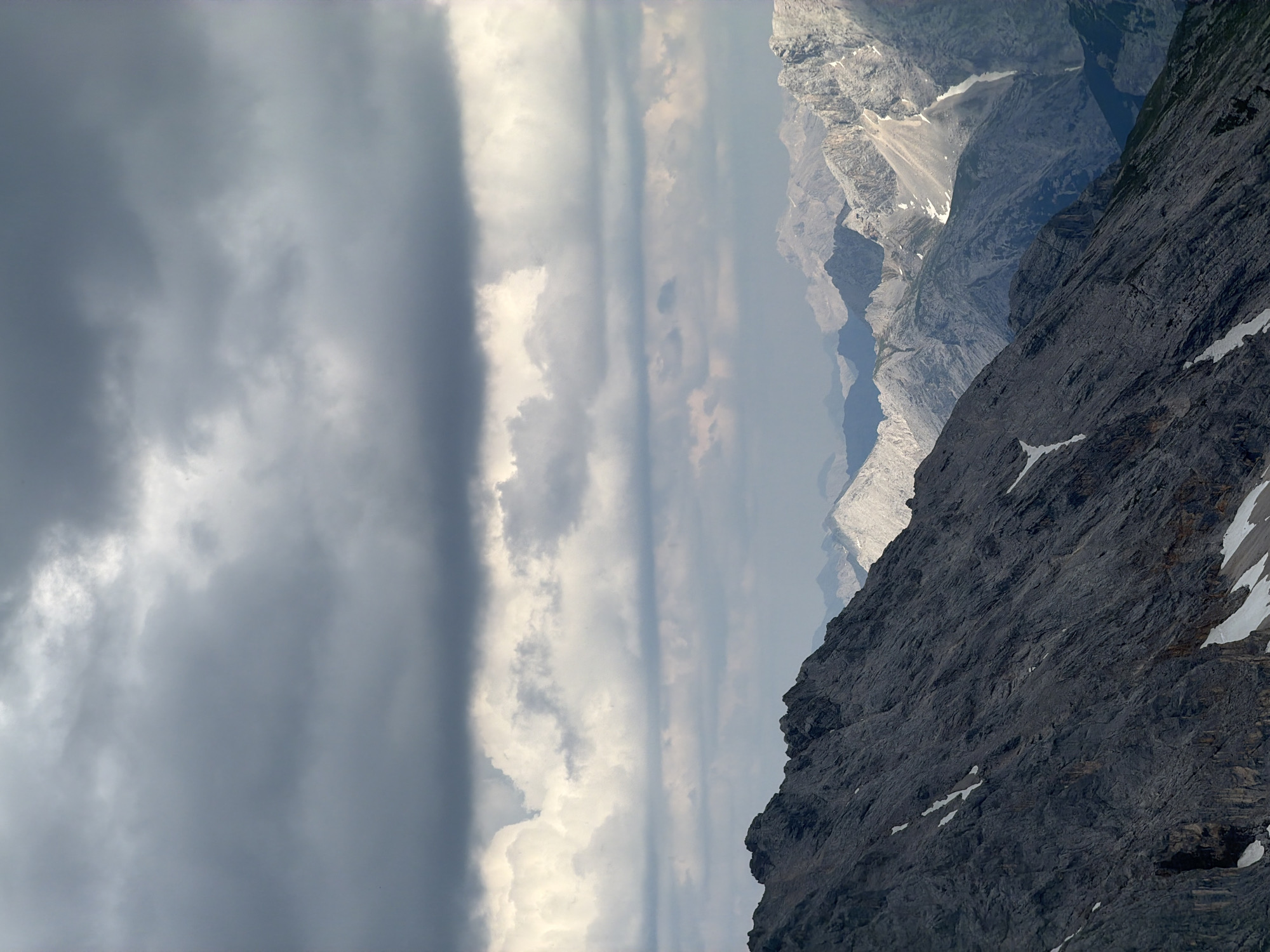 The image depicts a dramatic mountain landscape in Garmisch-Partenkirchen, Germany. The scene is dominated by steep, rocky mountain faces on the right side of the frame, with patches of snow visible on the higher elevations. The rock formations are dark and textured, suggesting a rugged and imposing terrain. In the background, a series of layered mountain ranges recede into a hazy, muted blue distance, indicating significant depth and scale.
The left side of the image is filled with a vast expanse of sky, characterized by a dramatic contrast between dark, heavy storm clouds on the far left and brighter, more diffused clouds towards the center. The transition between the dark and light clouds creates a striking visual effect, hinting at changing weather conditions.
There are no discernible objects or text within the image. The scale and ruggedness of the mountains, combined with the dynamic cloud formations, evoke a sense of awe and the power of nature. The lighting suggests it might be late afternoon or early evening, with the sun attempting to break through or having recently set, casting a soft glow on the more distant peaks. The overall atmosphere is one of grand, untamed wilderness. There are no visible people in the image.