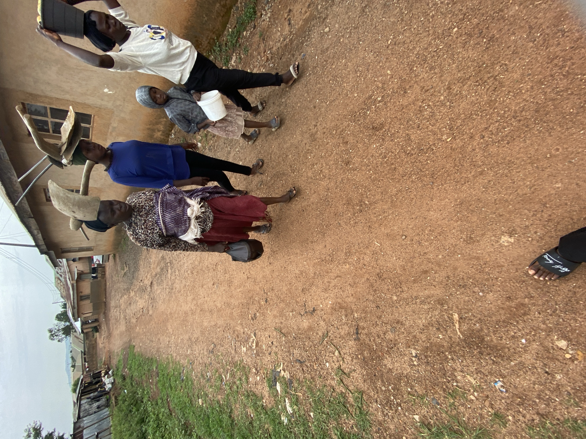 The image shows a dirt road in what appears to be a residential area of Bukuru, Nigeria. From left to right are buildings, including some tin shacks, and vegetation. Four people are on the road. A person in a white shirt and black pants holds a tablet and looks like they may be taking a photo. Another person wears a gray hoodie. Further down the road is a person wearing a blue shirt and black pants. Closer to the camera is a person wearing a horned headdress, patterned top, and red skirt.