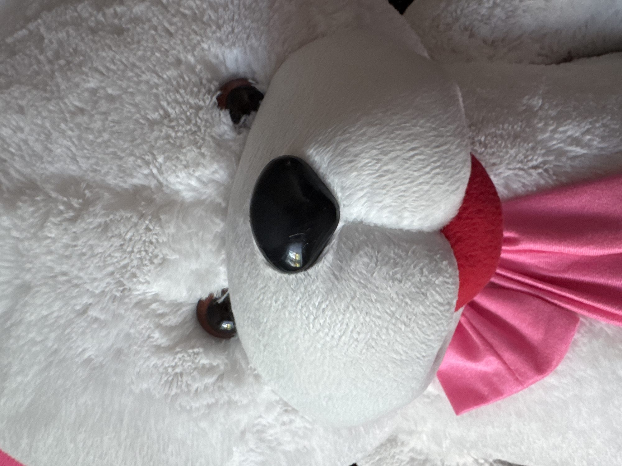 This is a close-up, head-on, full-frame shot of a white teddy bear. The teddy bear has large, dark, glossy eyes and a prominent black nose. It is wearing a pink ribbon tied into a bow around its neck. The fur of the teddy bear appears soft and fluffy. The background is blurred and out of focus. There is no visible indication of the location being Lysaya Gora, Russia.