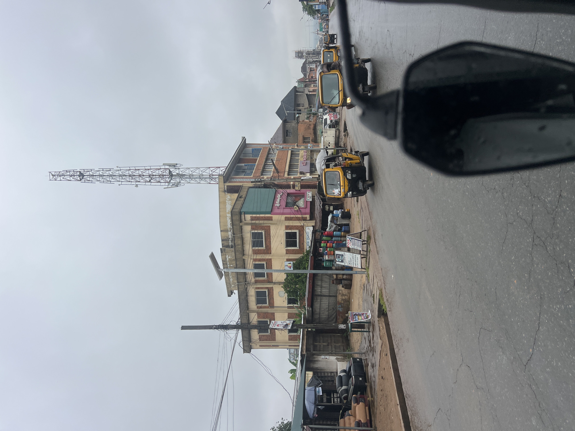 The image captures a street scene in Onitsha, Nigeria, under an overcast sky. A wide, wet asphalt road stretches into the distance, marked by cracks and puddles, suggesting recent rainfall. Several yellow auto-rickshaws, also known as "keke," navigate the road, some in motion and others parked. The buildings lining the street vary in architecture, with some featuring multiple stories and balconies, while others appear to be single-story shops. Prominently, a tall telecommunications tower rises above the cityscape. On the left side of the road, there is a shop with an umbrella providing shade over some furniture and stacked tires. Nearby, various items are displayed outside shops, including colorful plastic containers, a laptop, and advertising boards. A utility pole with numerous wires is visible near the center of the frame. The overall impression is of a bustling urban environment, likely during a day with poor weather conditions.