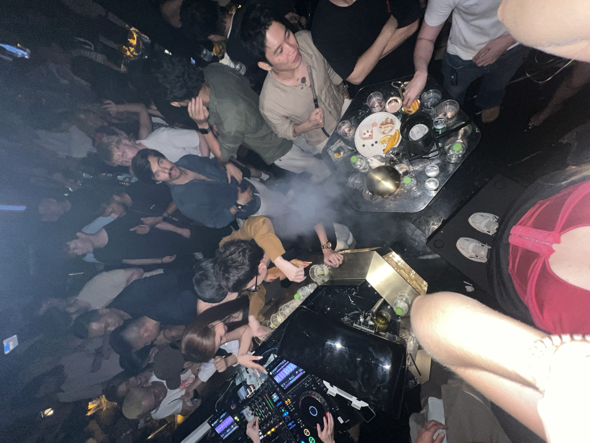 The image depicts a crowded indoor space, likely a nightclub or bar, in what appears to be the city of Thủ Đức, Vietnam. A large group of young people are present, mostly standing or leaning in. Visible are individuals with diverse clothing styles, ranging from casual t-shirts to collared shirts and dresses. Some people are holding drinks, and a few are clustered around tables.
A DJ booth is prominent in the foreground, equipped with DJ mixing equipment and people operating it. A table nearby contains food platters, drink glasses, water bottles, and a smoking apparatus, with people interacting around it. Smoke or vapor is visible in the air, possibly from the smoking equipment. The environment is darkly lit with artificial lighting that casts shadows.