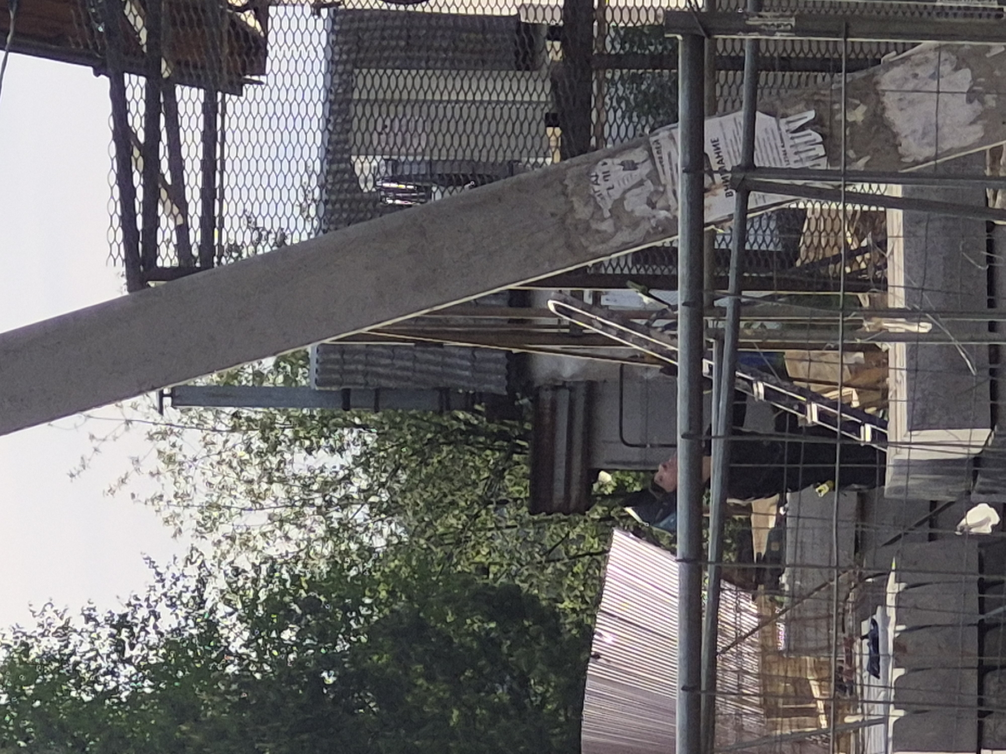An outdoor scene depicts a fenced-off area under what appears to be an elevated walkway or structure, viewed from an upward angle. The scene is complex with various elements overlapping.
On the lower left, there are trees with dense green foliage. Adjacent to the trees, a corrugated metal roof is visible. Behind the fence, a person wearing dark clothing seems to be on a ladder or scaffolding.
The metal fencing has a grid pattern, and there are additional metal supports and beams creating a cluttered foreground. A large, flat, concrete or stone beam extends diagonally across the image. Flyers or posters are adhered to some of the vertical metal beams. Above this are metal cages.
The overall setting suggests an urban environment in a state of construction or disrepair.
