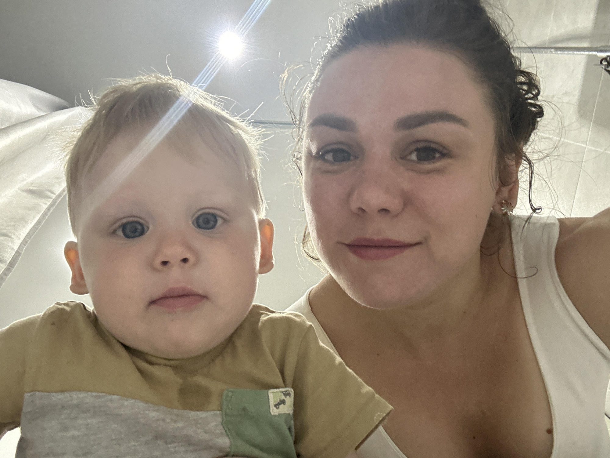The image captures an upward-angled close-up of an adult woman holding a baby. The woman, occupying the lower portion of the frame, has dark hair, brown eyes, and fair skin, with a slight smile on her pink lips. She wears a white tank top, exposing part of her chest with a visible mole and an earring on her left ear. The baby, positioned above and slightly to the right of the woman, has light blonde, fine hair, wide blue eyes, and pink lips, maintaining a neutral expression. The baby is dressed in a light tan short-sleeved top with a grey trim and a small green patch pocket on the left side bearing a white label. Both individuals are looking directly at the camera. The background consists of a uniformly bright, light-colored surface, possibly a wall or ceiling, with some subtle vertical textures on the upper left. A distinct, bright light source in the upper right corner produces a strong lens flare. The bottom right edge reveals white tiled surfaces and a partial view of a metallic fixture, suggesting an indoor setting, likely a bathroom. Based on the provided instruction, the scene, if located in Chaikovsky, Russia, depicts this interaction within that city.