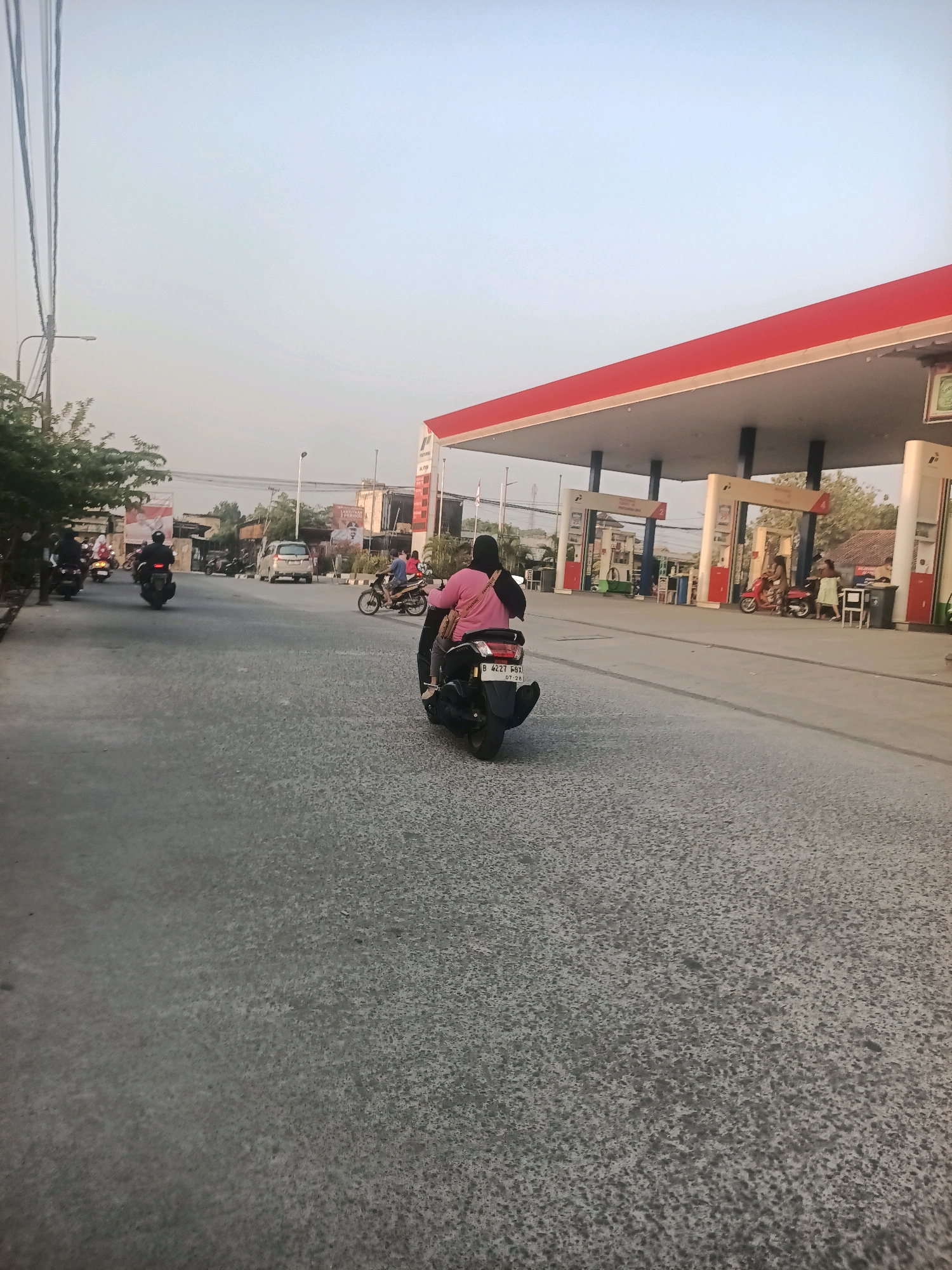 A street scene in Wanajaya, Indonesia, features a gas station with a red roof and white pillars. Several people are present, with a woman in a pink shirt riding a motorcycle in the foreground, her back to the camera. Another motorcycle with two riders is behind her, and more motorcycles are visible in the background. A silver car is driving on the road. The sky is pale blue. The road surface is grey and textured.
