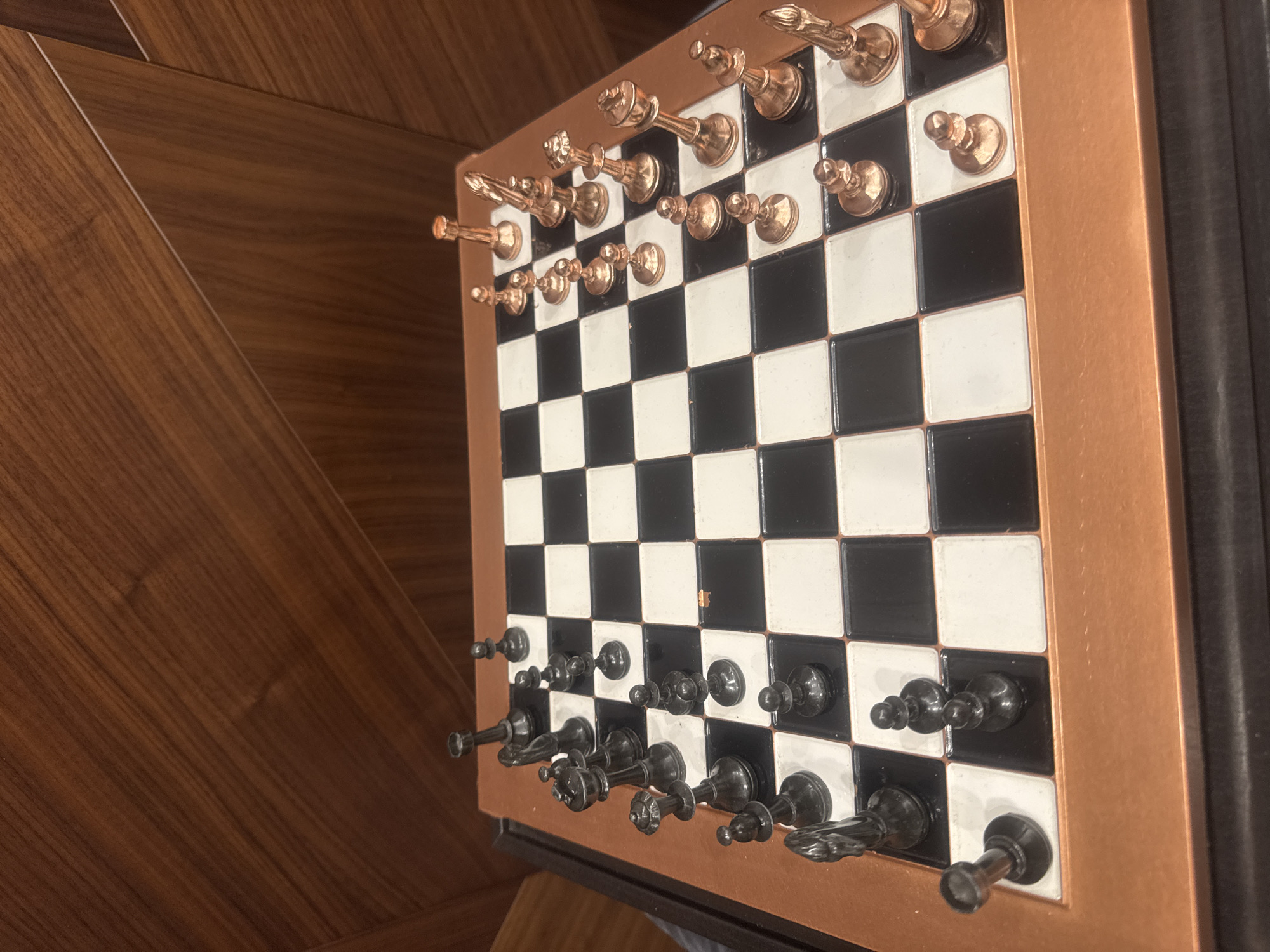 The image shows a chessboard with pieces set up for a game. The board has black and white squares and a copper-colored frame. The chess pieces are in two colors: copper and black. They are ornate and appear to be made of metal. The board and pieces sit on a surface that appears to be made of wood with a diagonal pattern. The chessboard leans against a darker, possibly black, vertical surface. The scene's location in Yerevan, Armenia is not apparent from the visual data.