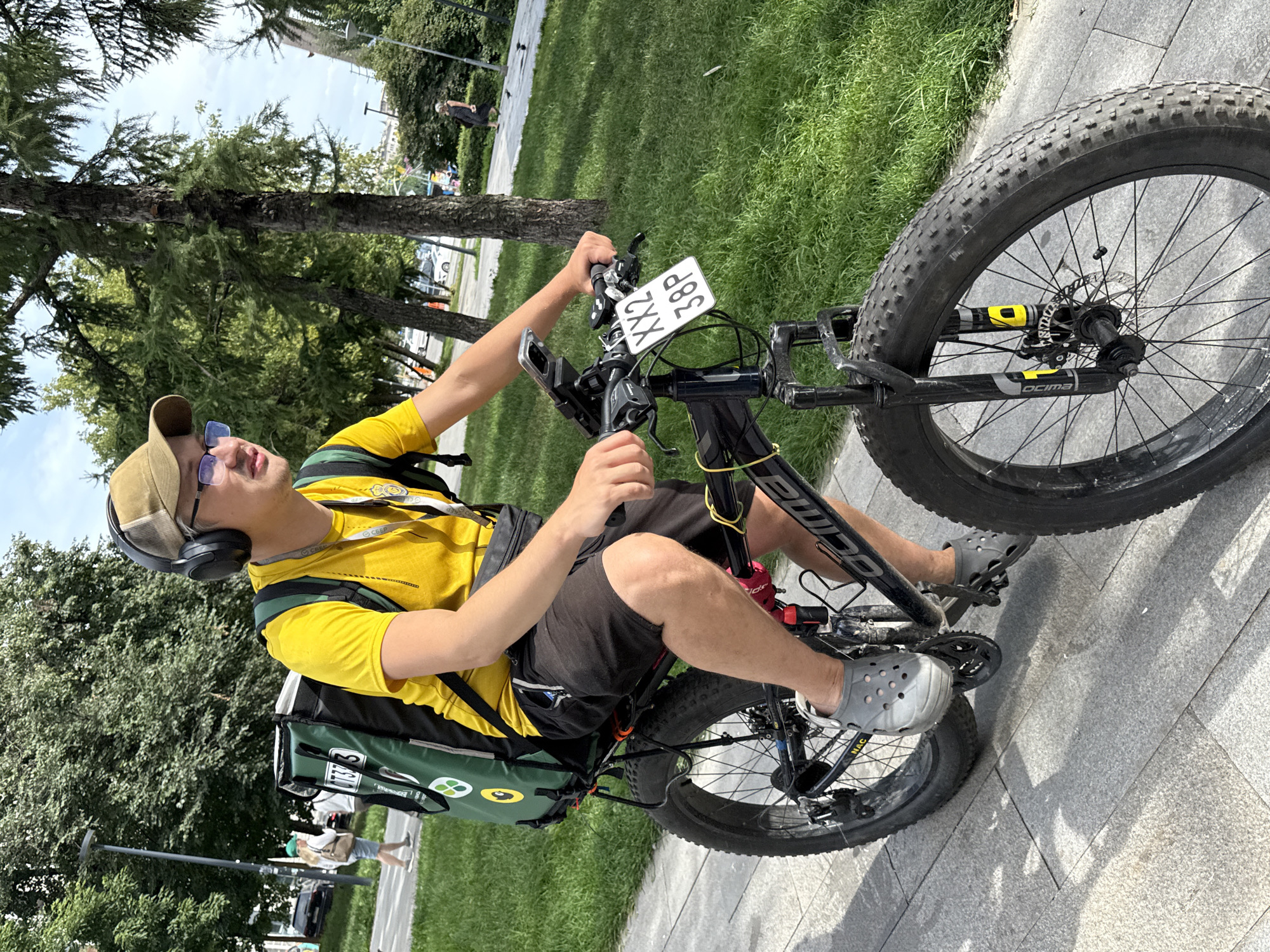 An individual wearing a yellow t-shirt, black shorts, gray Crocs, a beige baseball cap, and glasses rides a fat-tire bicycle on a paved path in a park-like setting. The person has headphones on and is looking to the right. A large green delivery backpack is mounted on a rack behind the bicycle's seat. A white sign with black lettering "XX2 38P" is attached to the handlebars. Trees and grass are visible in the background, along with some parked cars on a street in the distance. The sky is partly cloudy. The scene appears to be outdoors in a city, likely Moscow, Russia given the context, during daylight hours.