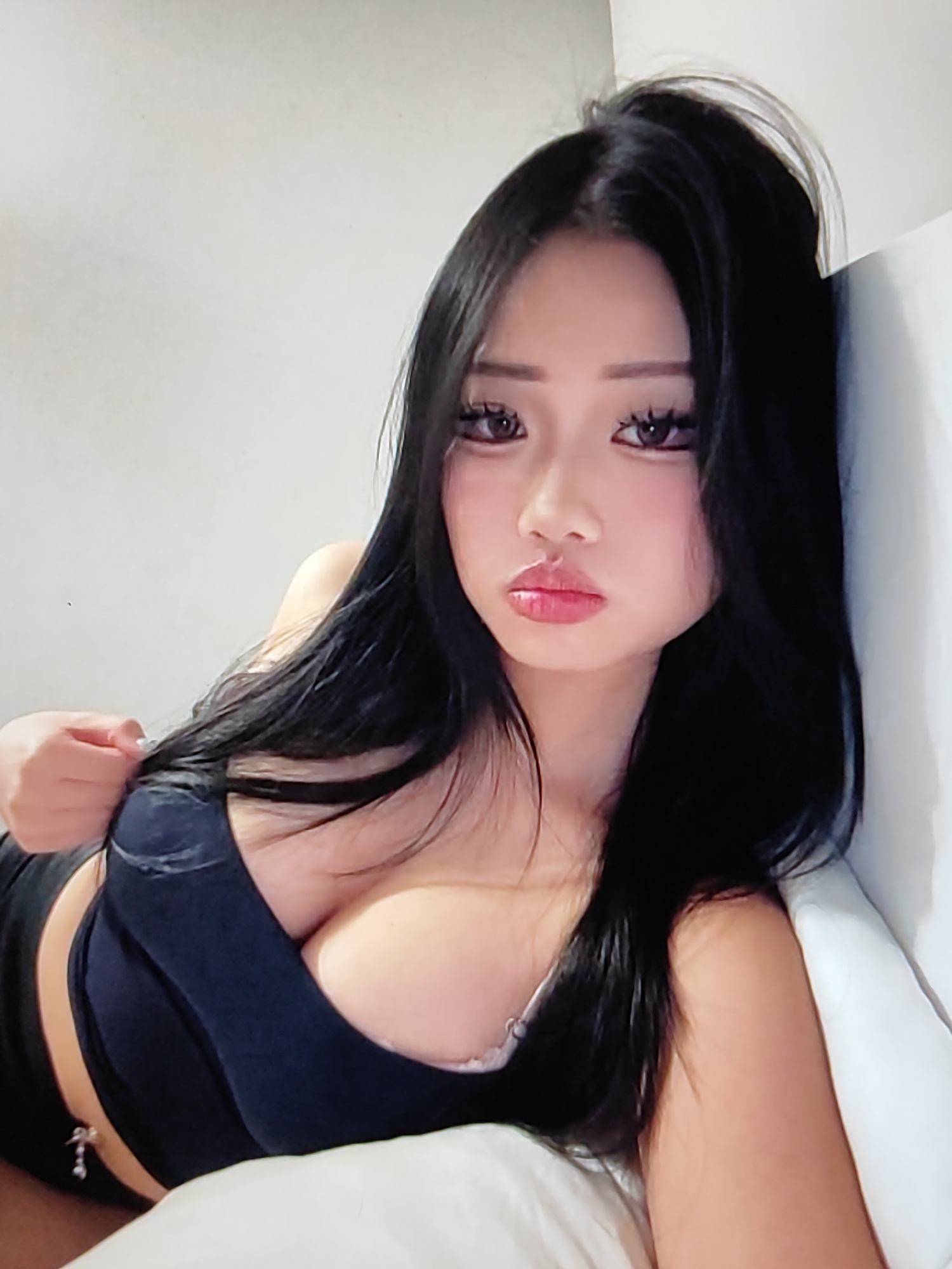 The image shows a close-up of a young woman with long, black hair, lying on a bed. She is wearing a dark-colored top that reveals her cleavage. Her eyes are dark and her lips are full and painted with pink lipstick. Her gaze is directed towards the camera. The background appears to be a plain, light-colored wall and a white pillow. There is no indication of the city of Fergana, Uzbekistan, in the image.