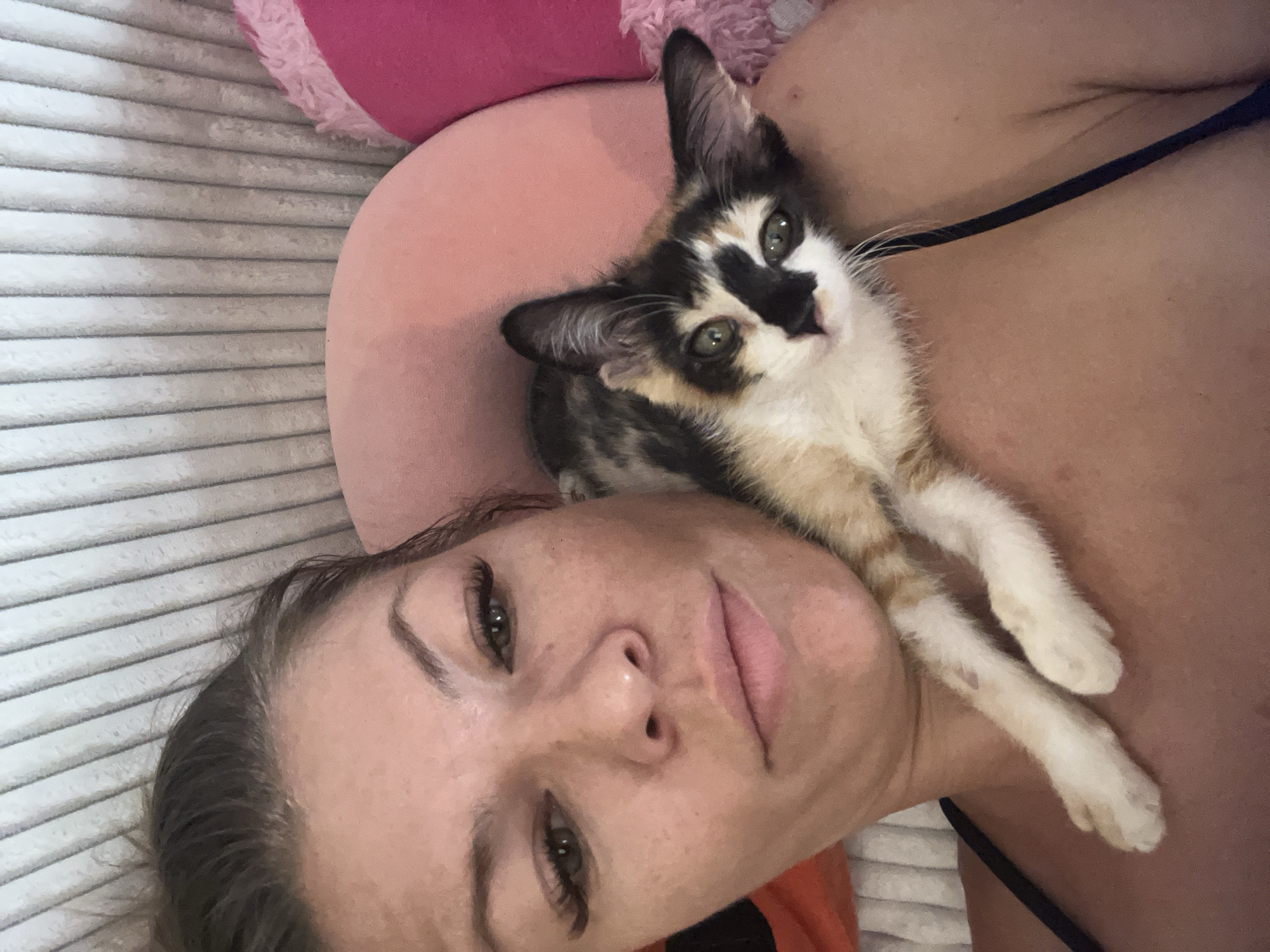 A woman is lying on a striped surface with a kitten on her chest. The kitten is calico-colored with white paws and a black and white face. The woman has light skin, dark hair, and green eyes. A pink pillow is visible in the background. The location in Kuta, Indonesia, is not discernible from the visual information provided.