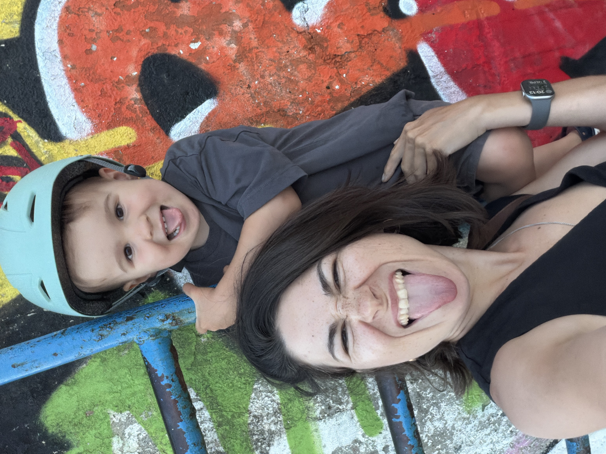 The image captures a playful selfie featuring an adult and a child lying down on a vibrant, graffiti-covered surface. Both individuals are looking up at the camera with wide smiles and their tongues sticking out, conveying a sense of fun and joy.
The adult, likely female, has dark hair and visible freckles on her nose and cheeks. She is wearing a black top and a thin silver necklace. On her left wrist, an Apple Watch is visible, displaying the time "20:13" (8:13 PM) and the date "2ЧЕРВ" (June 2nd, in Cyrillic).
Beside the adult is a young child wearing a light blue or mint green helmet and a dark gray t-shirt. The child mirrors the adult's expression, sticking out their tongue playfully and smiling.
The background is a striking canvas of colorful graffiti, featuring bold streaks and shapes in red, orange, yellow, green, black, and white, indicating an urban or artistic outdoor setting. A blue metal bar or railing, showing signs of wear and rust, runs across the lower part of the frame. The scene suggests a relaxed and happy moment, possibly during a late afternoon or early evening outing given the smartwatch time. The location context is Budva, Montenegro.