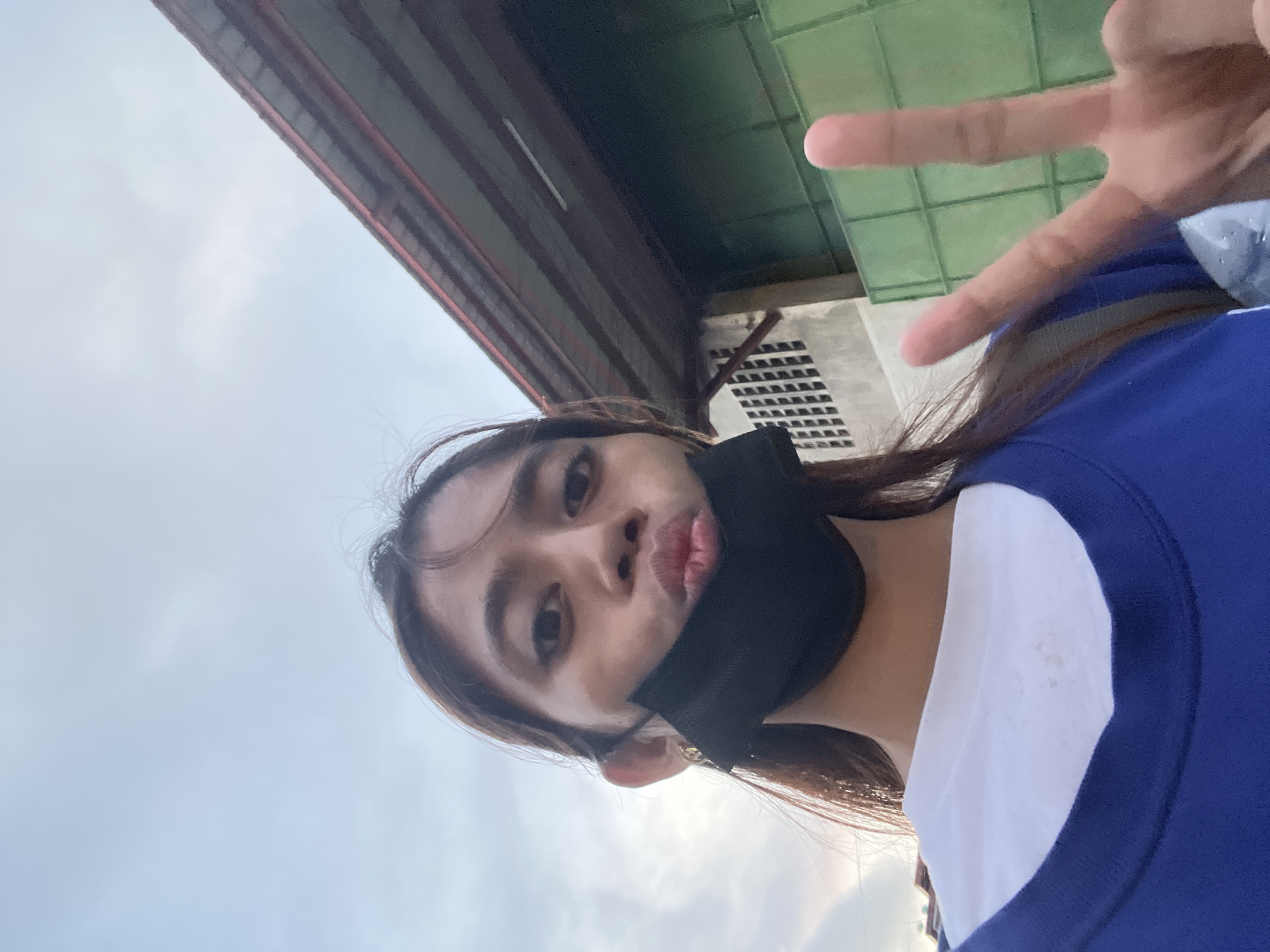 A young woman wearing a black face mask and a blue and white shirt is taking a selfie outdoors. She is making a peace sign with her right hand. In the background, there is a building with a corrugated metal roof and a green fence. The sky is cloudy. The location is likely an urban area in Davao City, Philippines, suggested by the building architecture and the prevalence of outdoor selfies in such settings.