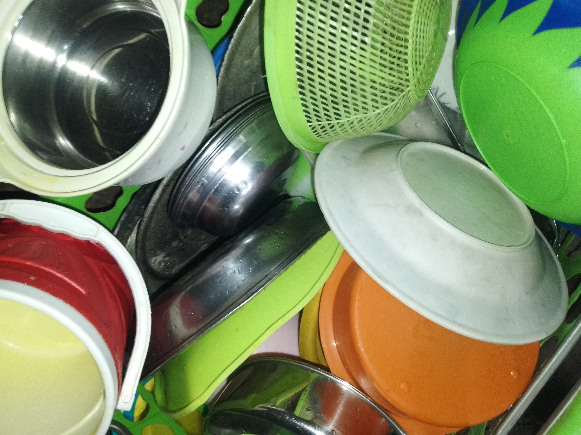 In a cluttered scene within Serti, Nigeria, a collection of kitchenware is piled together. The items include a stainless steel pot with a white lid, a green colander, a bright green bowl with a spiky pattern, a white plate, an orange plate, a red and yellow bucket, and several other metallic bowls and green plastic items. The objects appear to be wet, with water droplets visible on their surfaces, suggesting they may have recently been washed or are being stored in a damp environment. The overall impression is one of disarray and everyday domestic life in Serti.