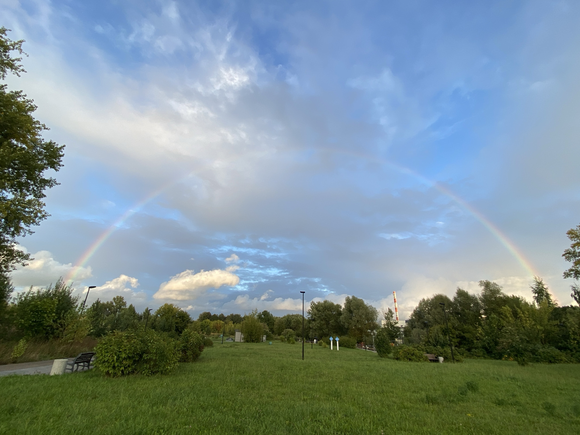 A wide-angle shot captures a park with a double rainbow arching across a blue and cloudy sky. The foreground is dominated by a lush green lawn, with a paved path and a bench on the left. In the middle ground, a line of trees and bushes separates the lawn from a distant industrial area. The industrial area is characterized by a tall, red and white striped smokestack. The sky is a mix of bright blue and soft, white clouds, with the rainbow prominently displayed. There are no people or animals visible in the scene. The overall impression is one of natural beauty juxtaposed with industrial elements.