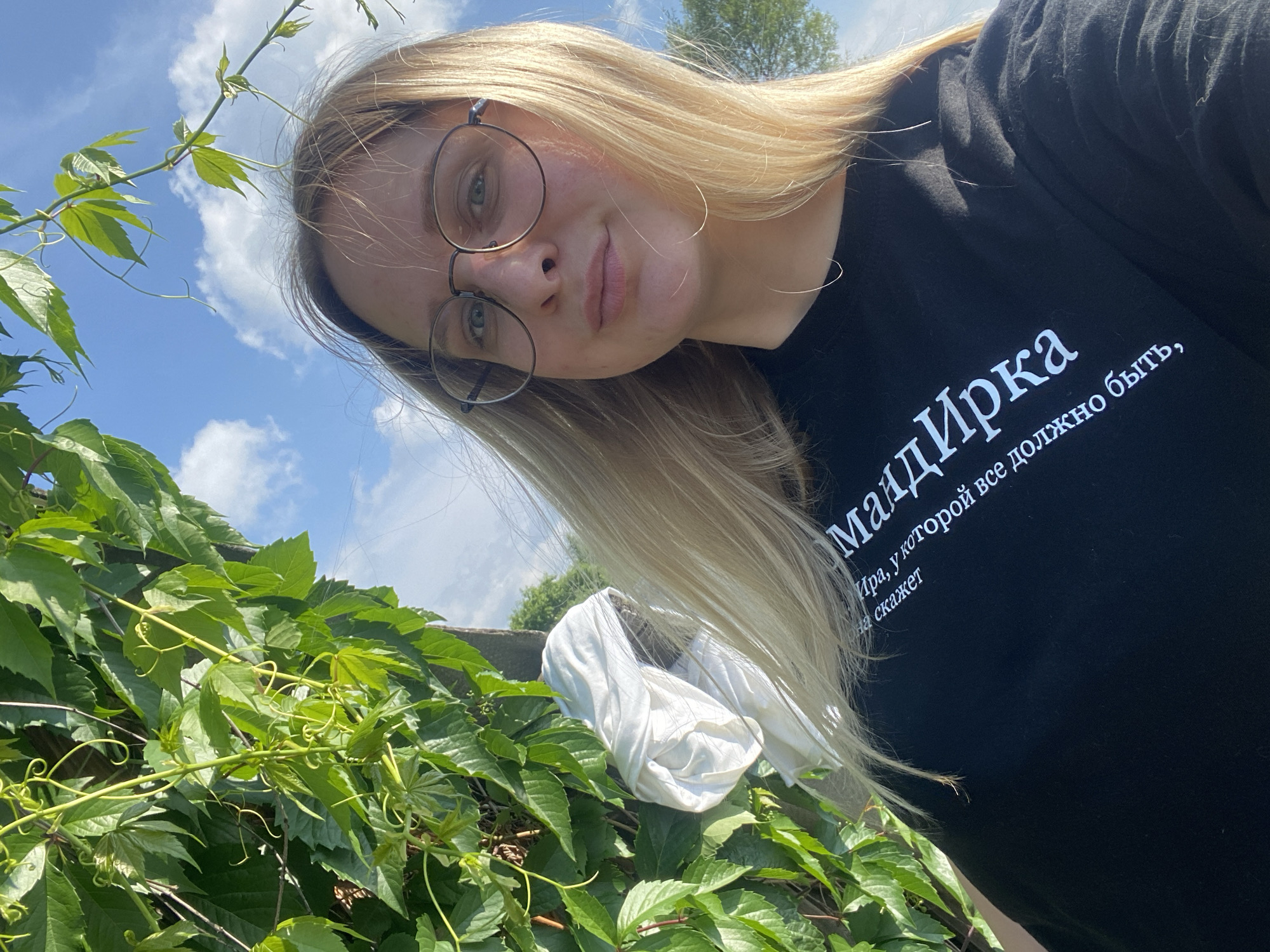 A young Caucasian woman with blonde hair and glasses is posing for a selfie outdoors. She is wearing a black t-shirt with white text on it. To her left, there is lush green foliage from a climbing plant, and further left, a white cloth is draped over what appears to be a pole or support. The background shows a bright blue sky with scattered clouds and some trees in the distance. The image was taken in the city of Kauchuk Stantsiya, Russia.