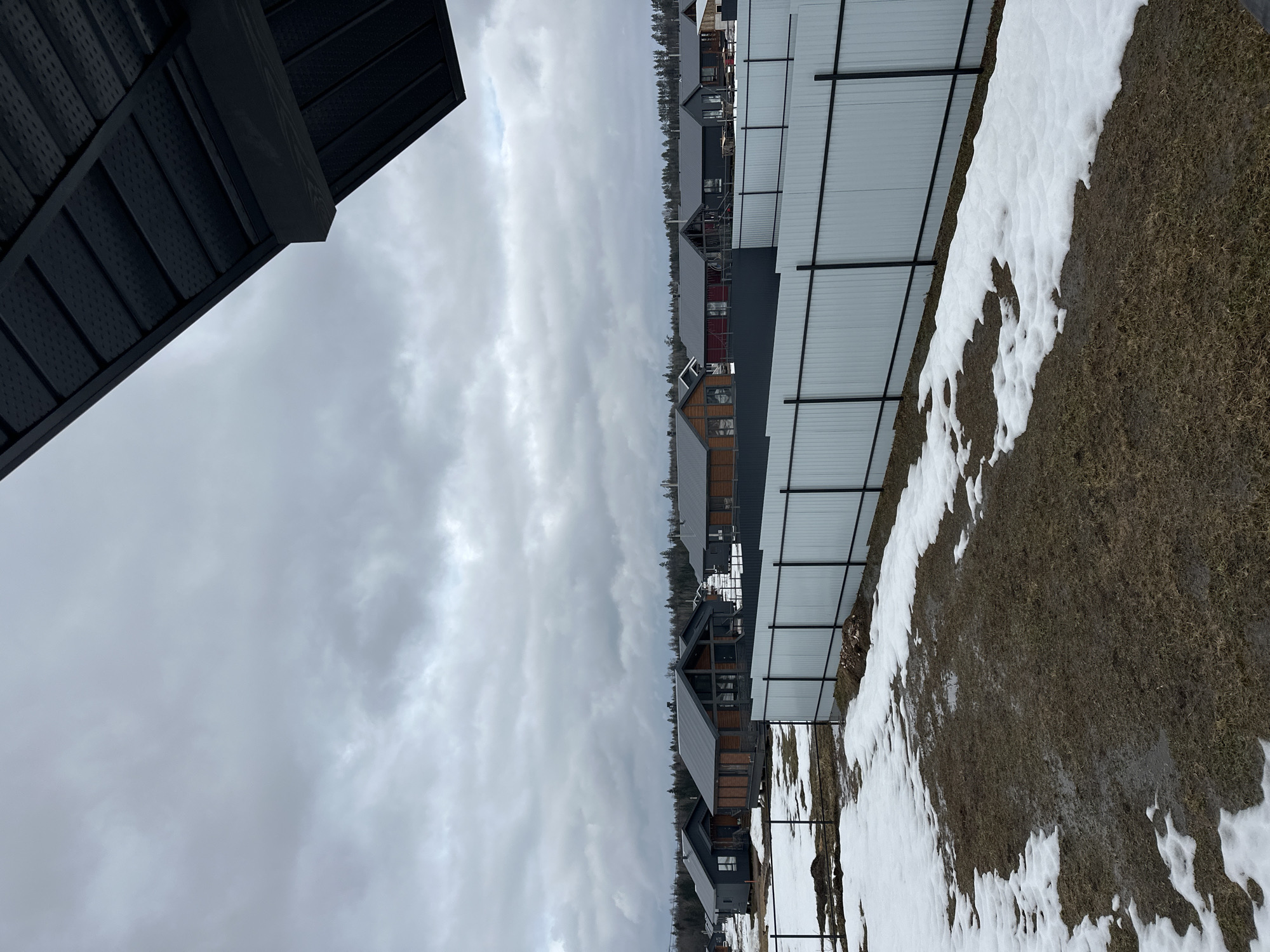 The image shows a row of modern houses under a cloudy sky. Snow is melting on the ground, revealing patches of brown grass. A fence runs along the side of the properties. The location is stated to be Подшивалово, Russia.